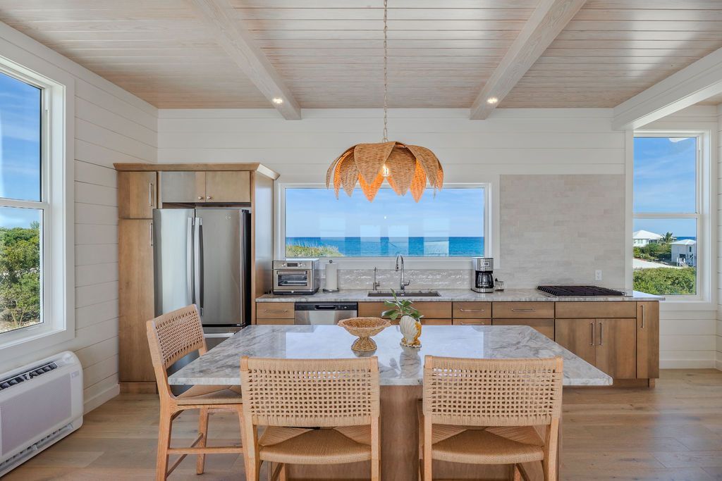 Bright coastal kitchen-dining room with woven chairs, wood island, and ocean view through large windows