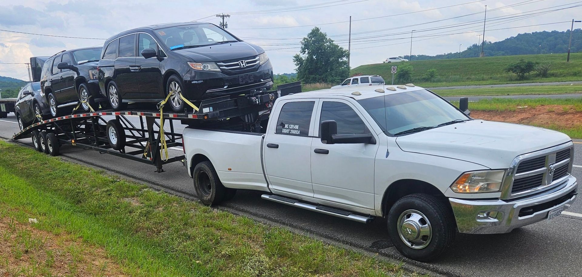 White tow truck hauling a black car on a trailer along a wet roadside under cloudy skies