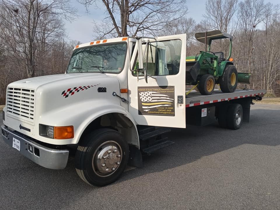 White flatbed tow truck carrying a green tractor on a road, with trees in the background