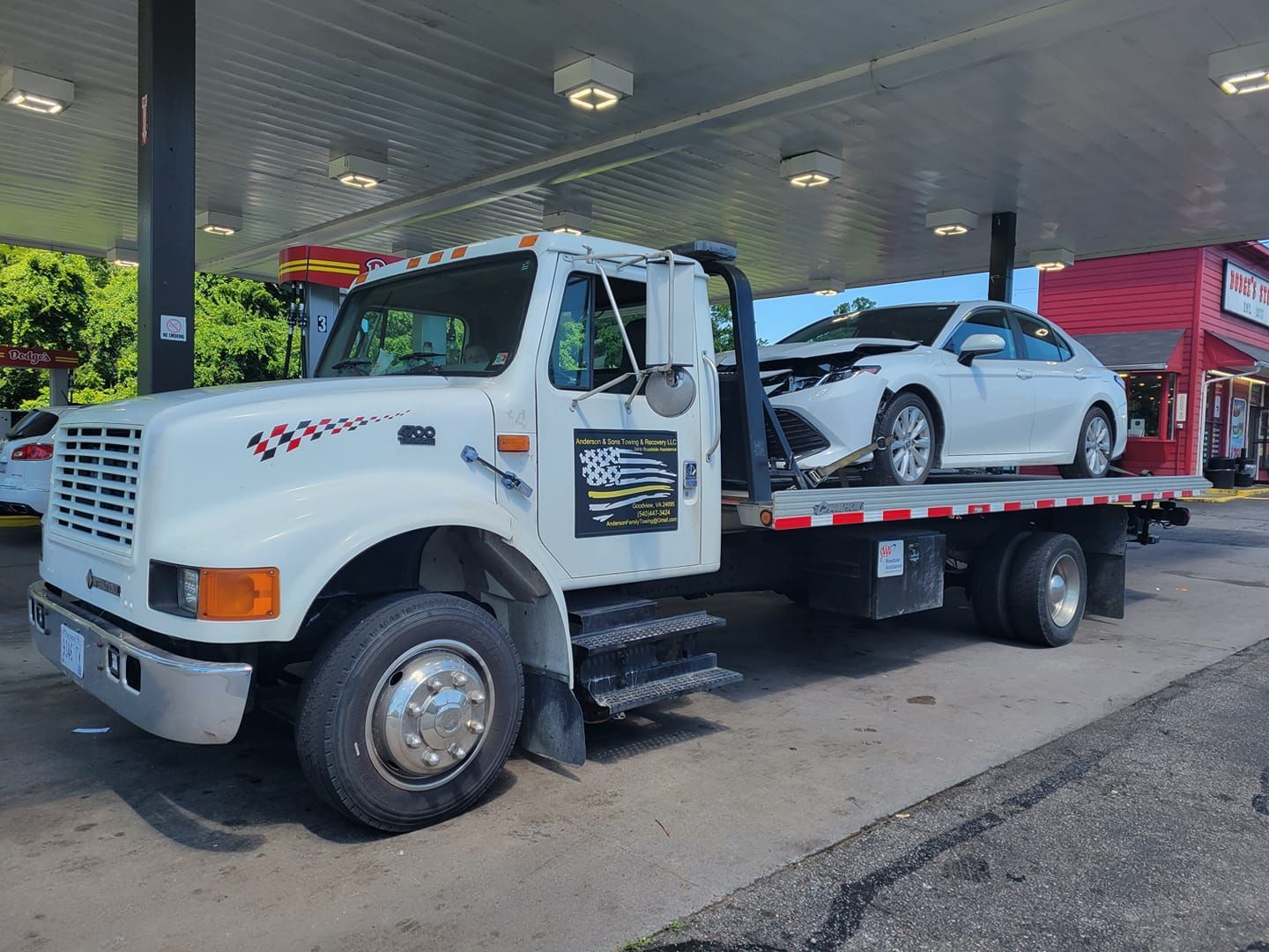 White tow truck carrying a white sports car at a gas station, with red canopy and pumps nearby