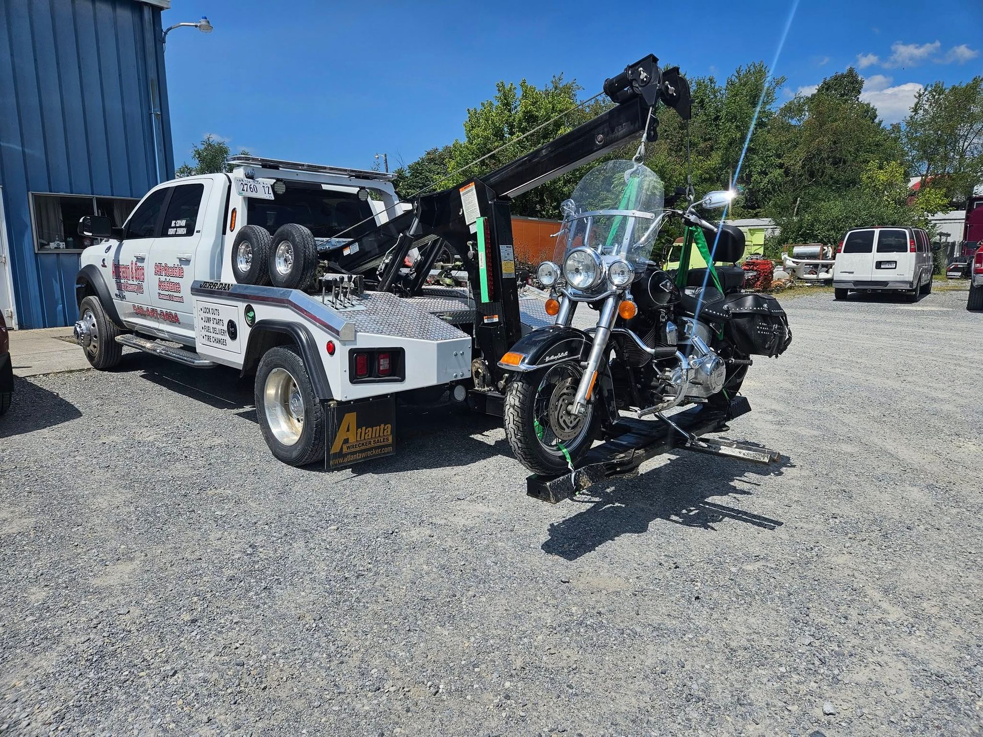 Tow truck lifting a damaged white SUV in a gravel lot, with workers nearby and a blue building behind.