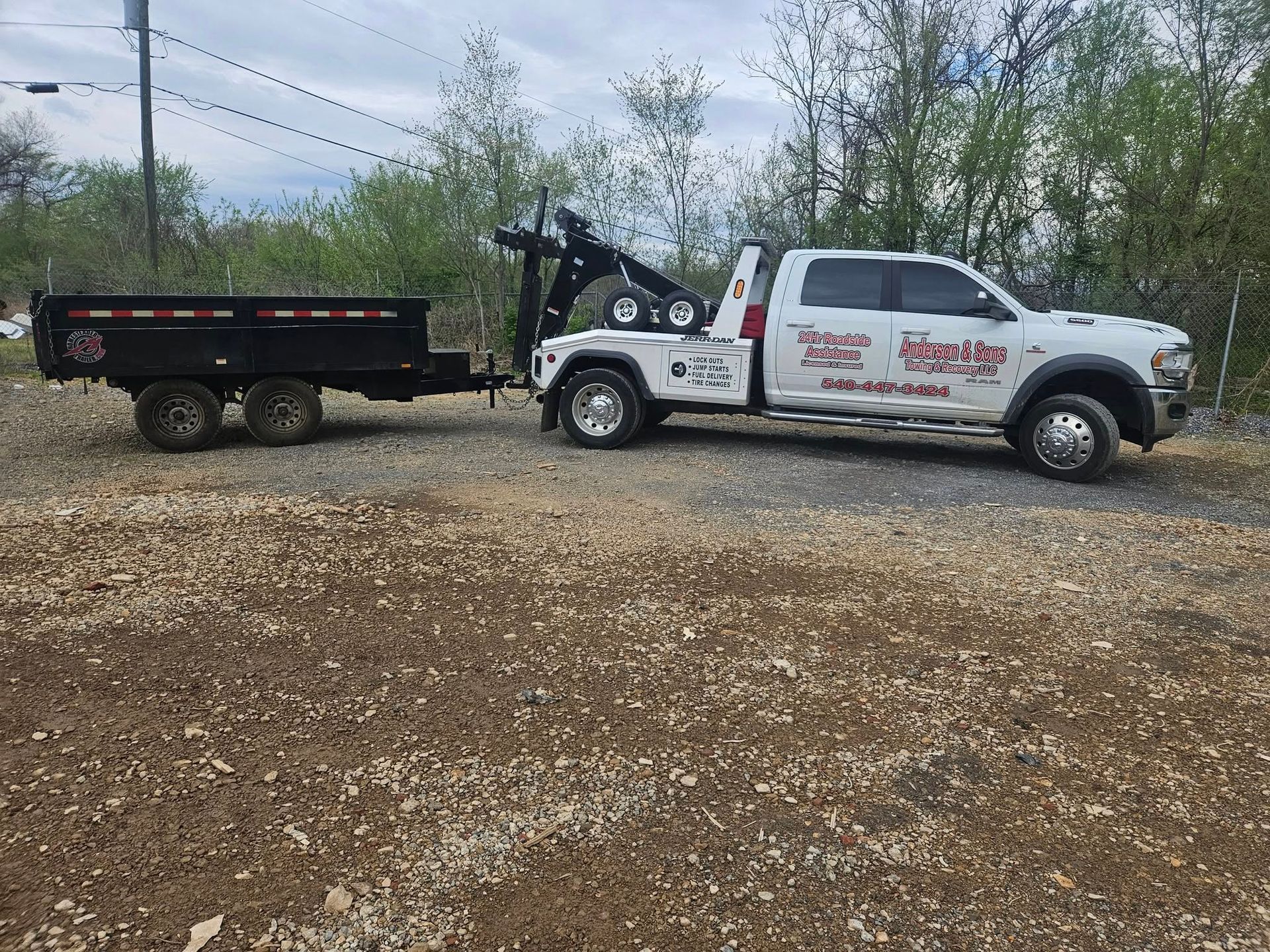 Tow truck with black trailer on gravel road beside trees and cloudy sky