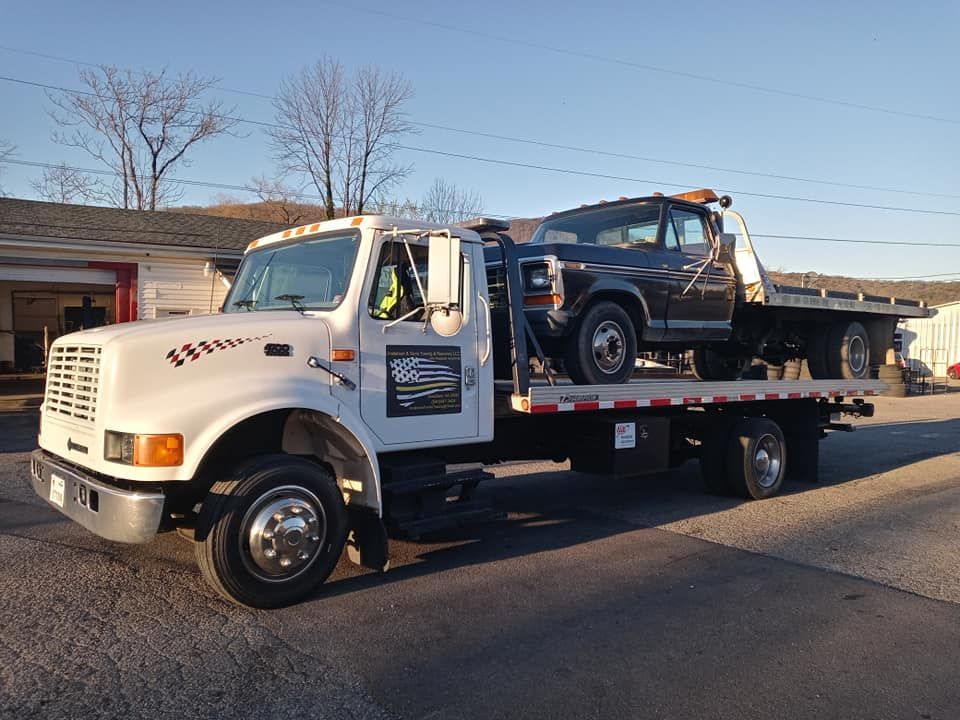 White flatbed tow truck carrying a dark blue SUV on a roadside at sunset