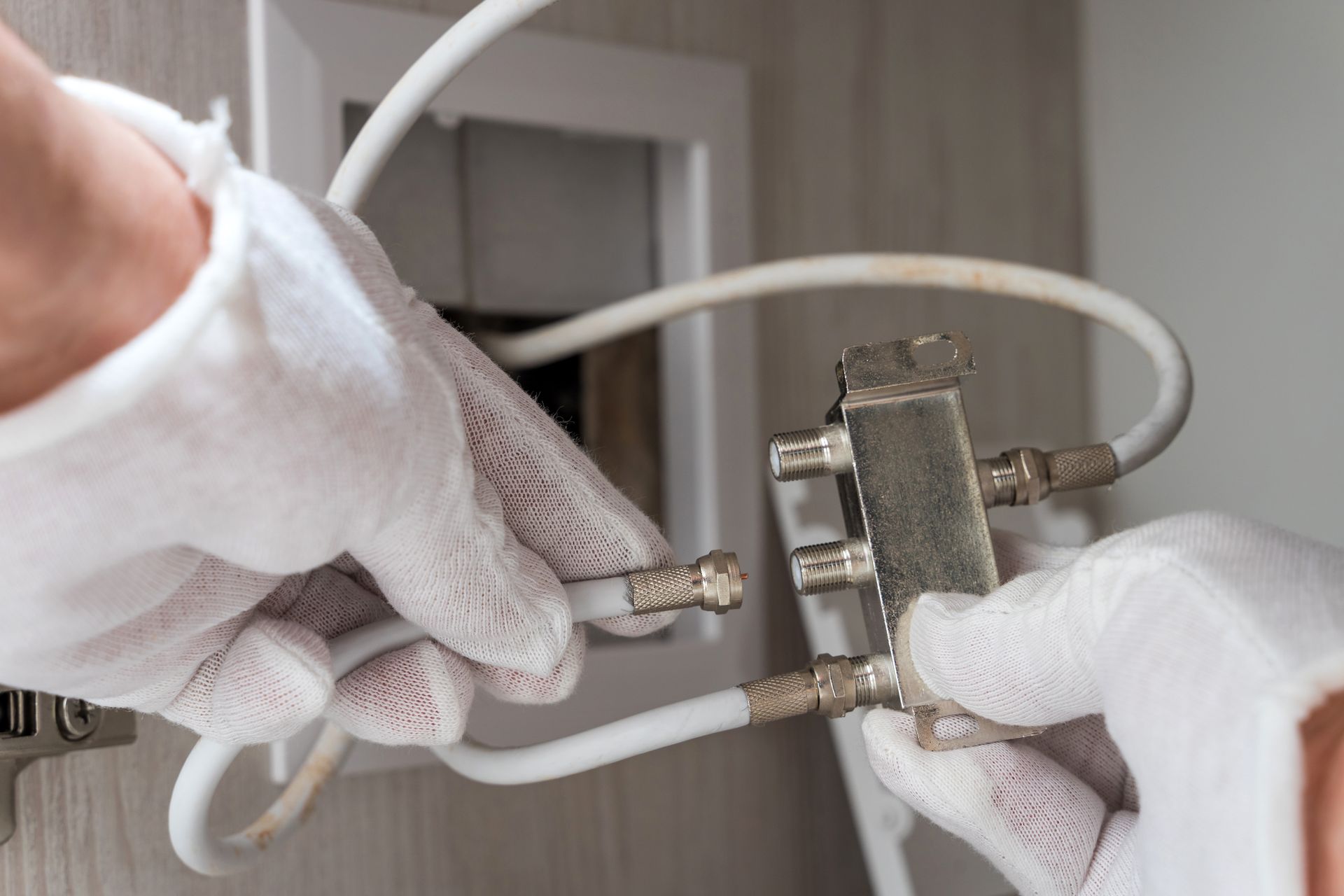 Gloved hands connecting a white cable to a wall-mounted port in a room
