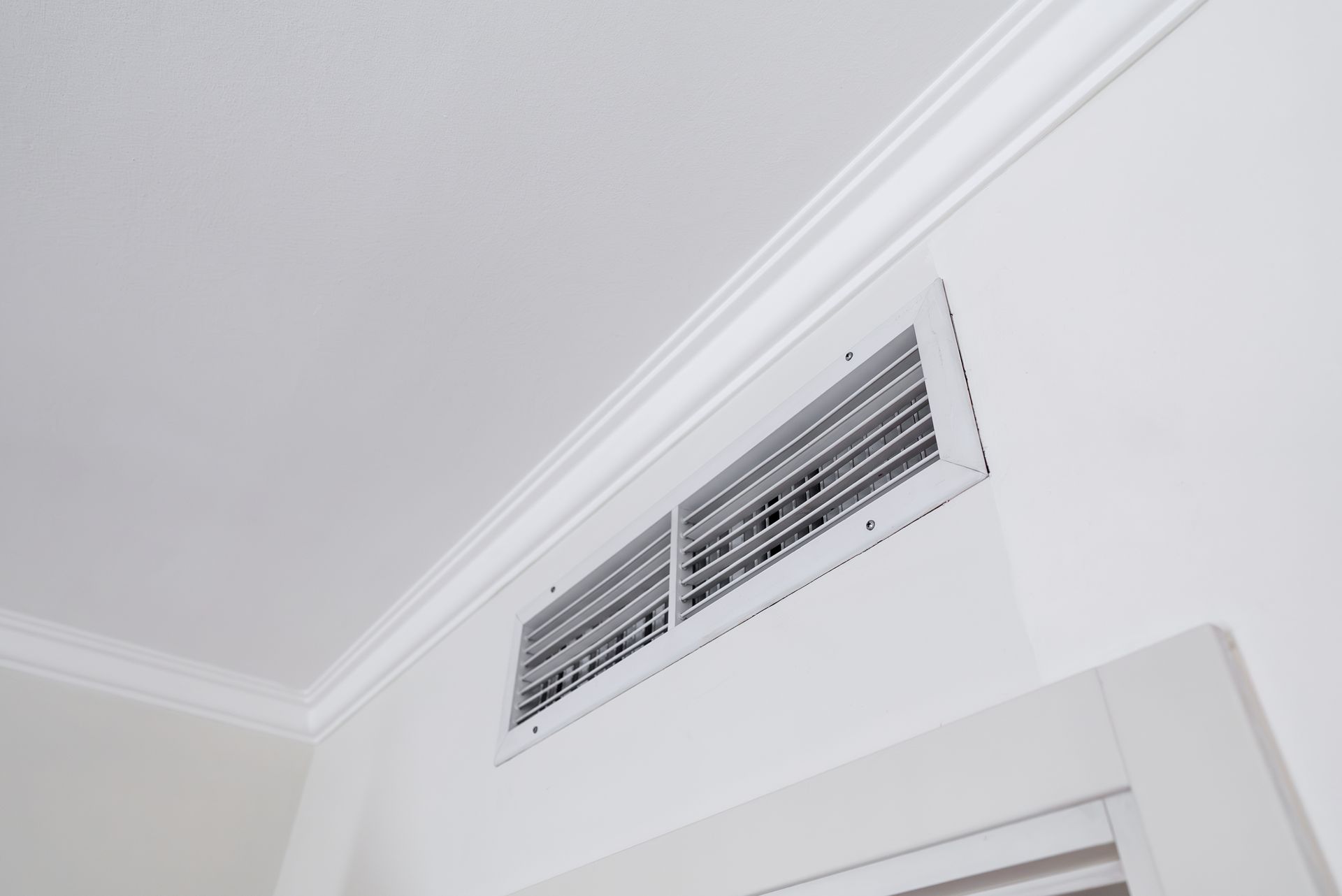 Wall vent near the ceiling above a door, painted white against a white wall and crown molding