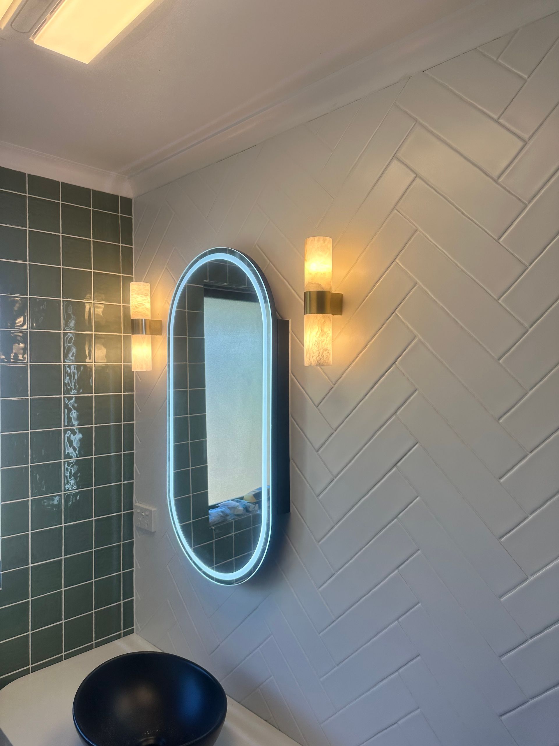 Modern bathroom with illuminated oval mirror, wall sconces, herringbone tile, and a black basin sink. Modern bathroom with illuminated oval mirror, wall sconces, herringbone tile, and a black basin sink.