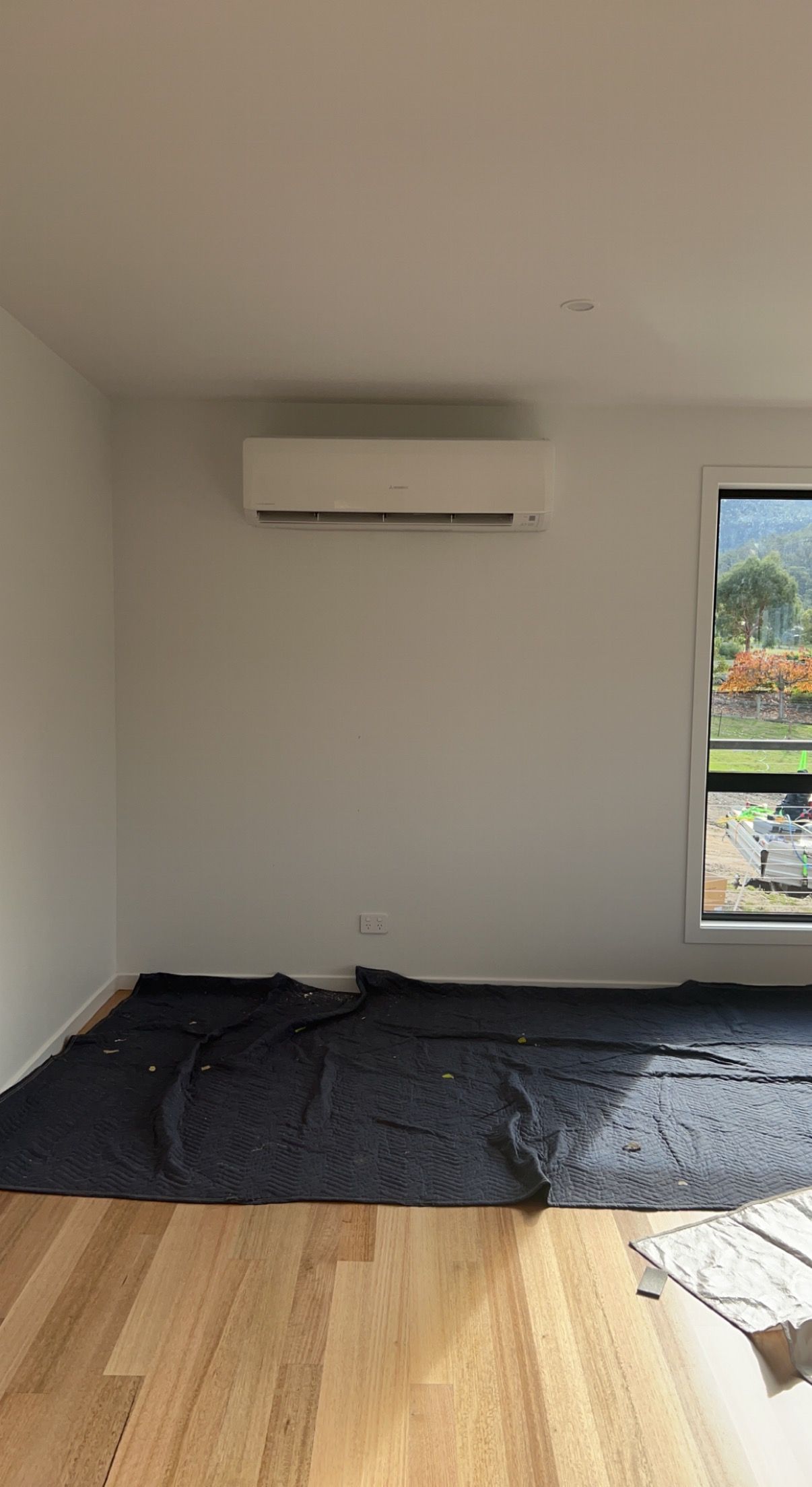 Empty room with wall-mounted AC, hardwood floor, dark tarp on floor, and window with greenery outside