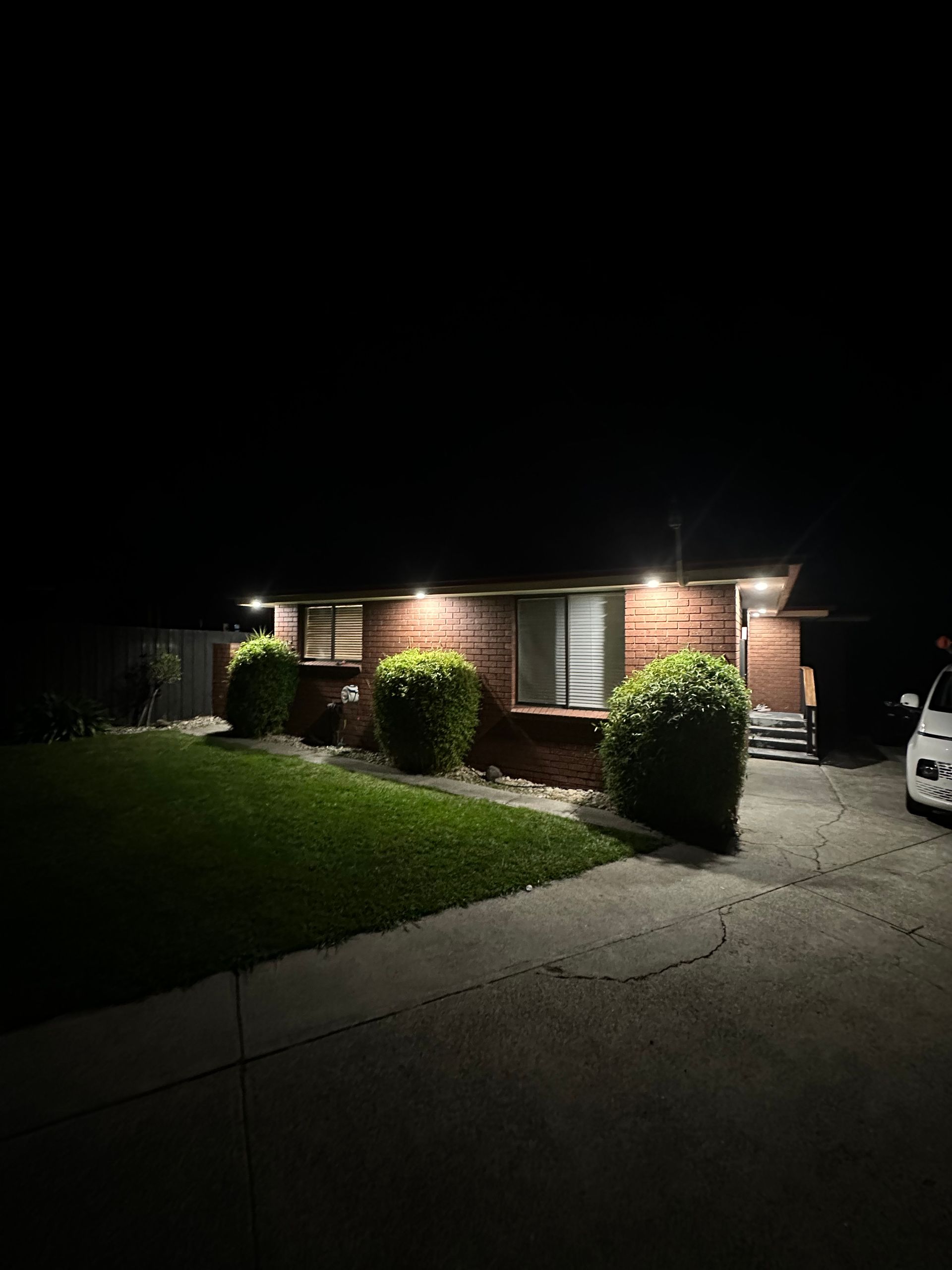 Nighttime view of a brick house with porch lights, trimmed bushes, lawn, and a driveway with a car at the edge
