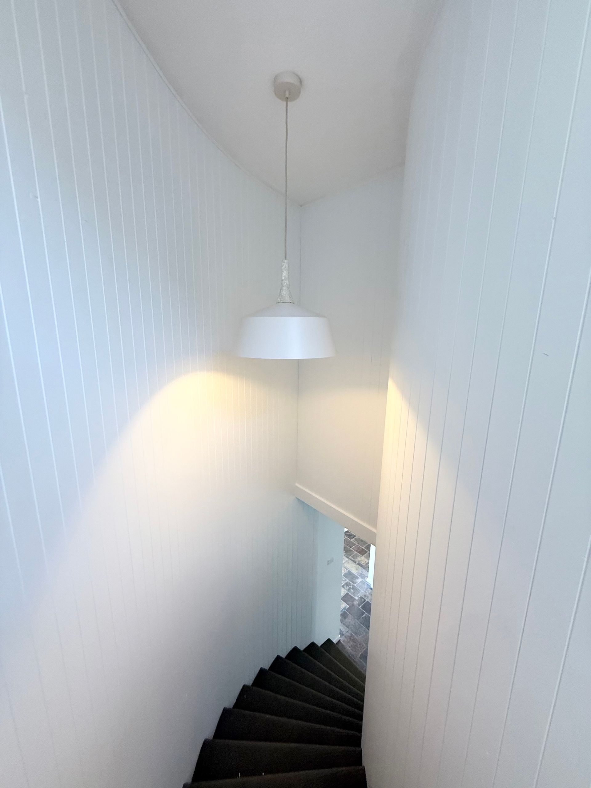 Staircase with dark steps descending in a narrow white hallway, lit by a hanging lamp.