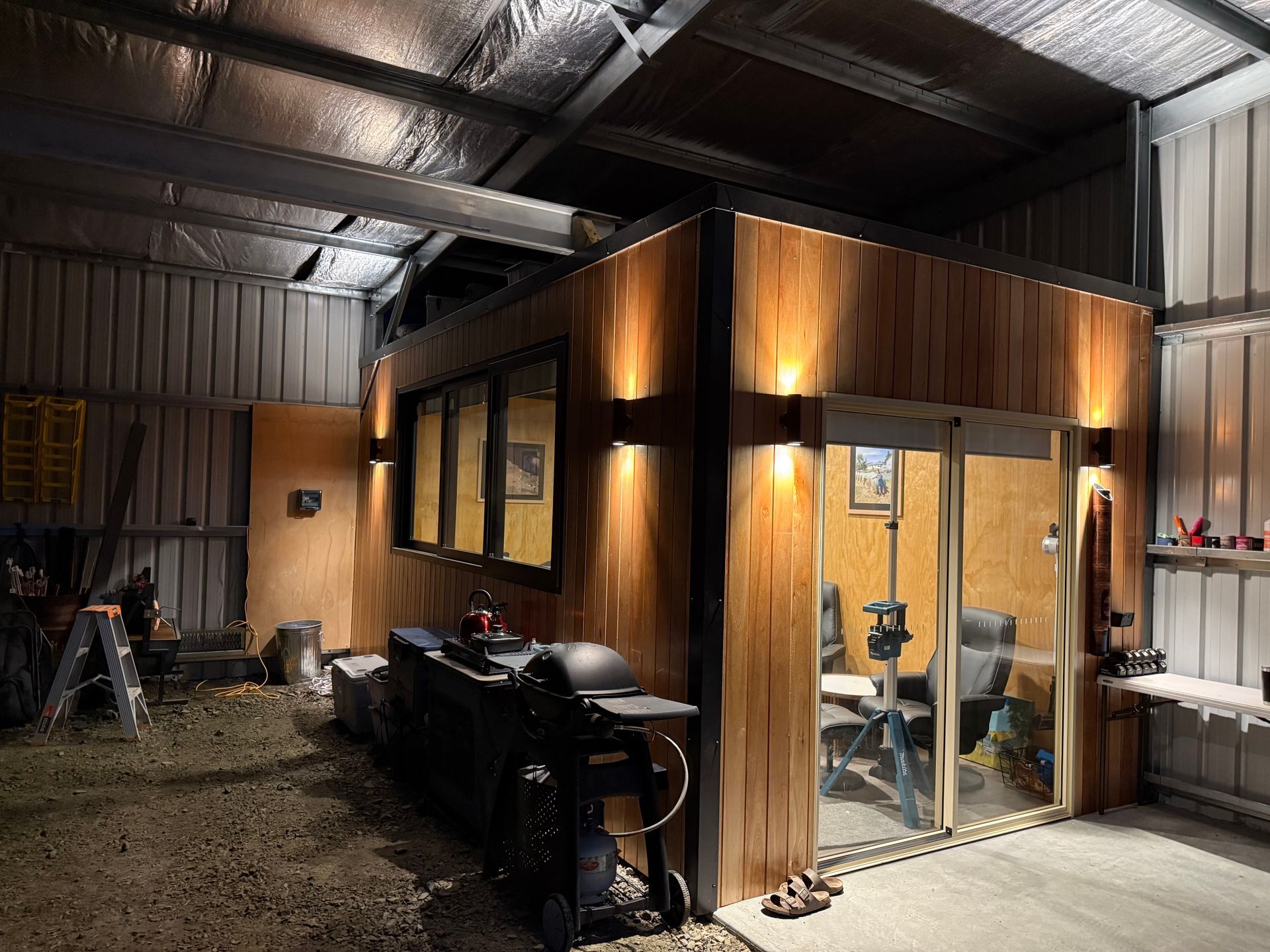Small wooden cabin inside a metal workshop, with glass doors, windows, and a generator outside.