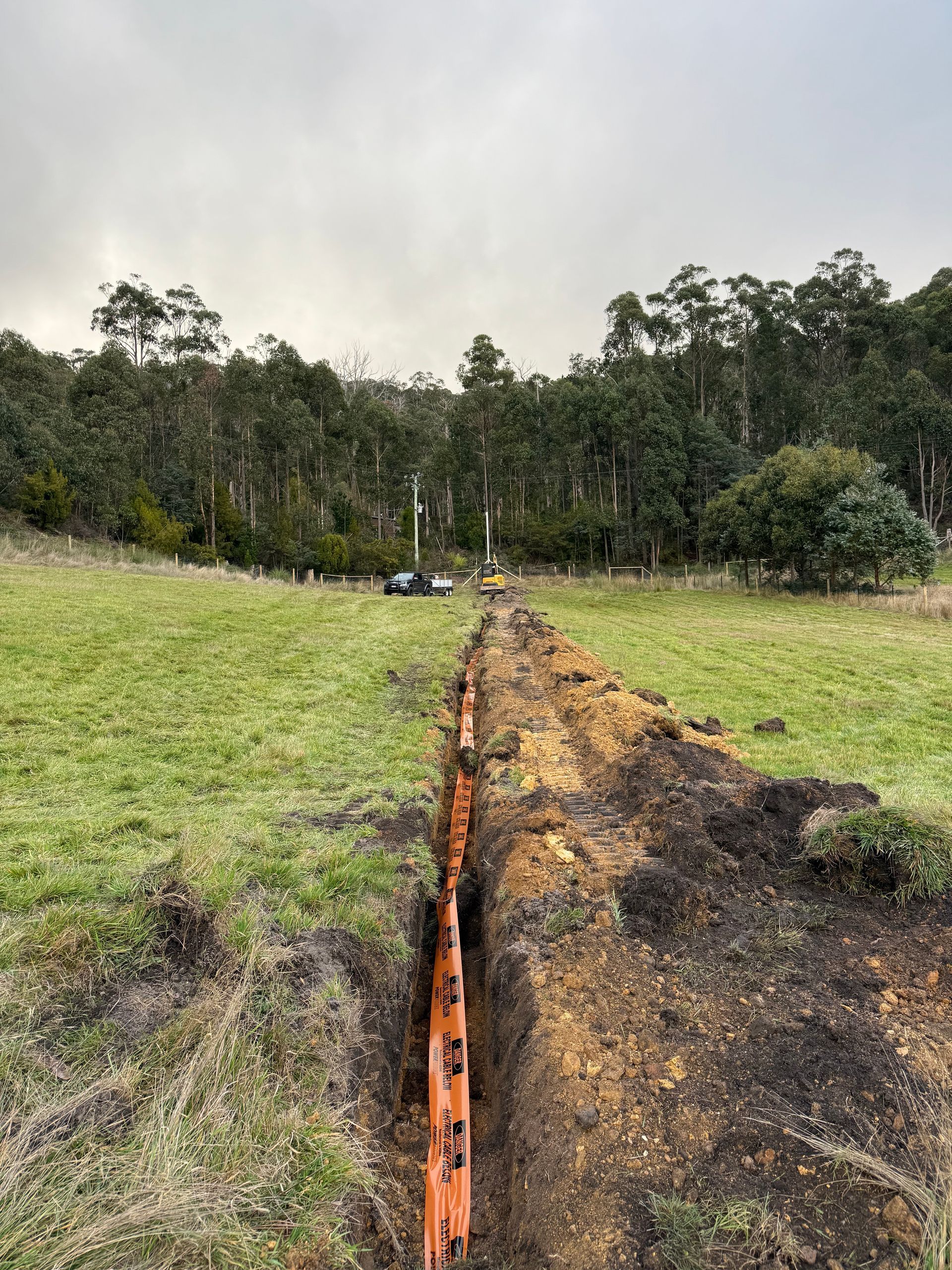 Narrow orange trench cutting through a grassy field toward a wooded hillside under an overcast sky