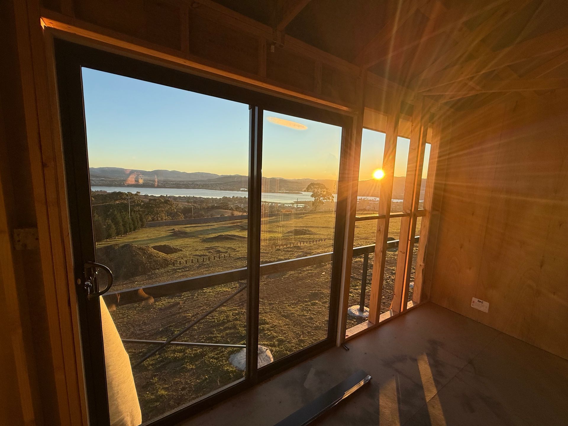 Sunset view through large windows in an unfinished room, with ocean and hills outside.