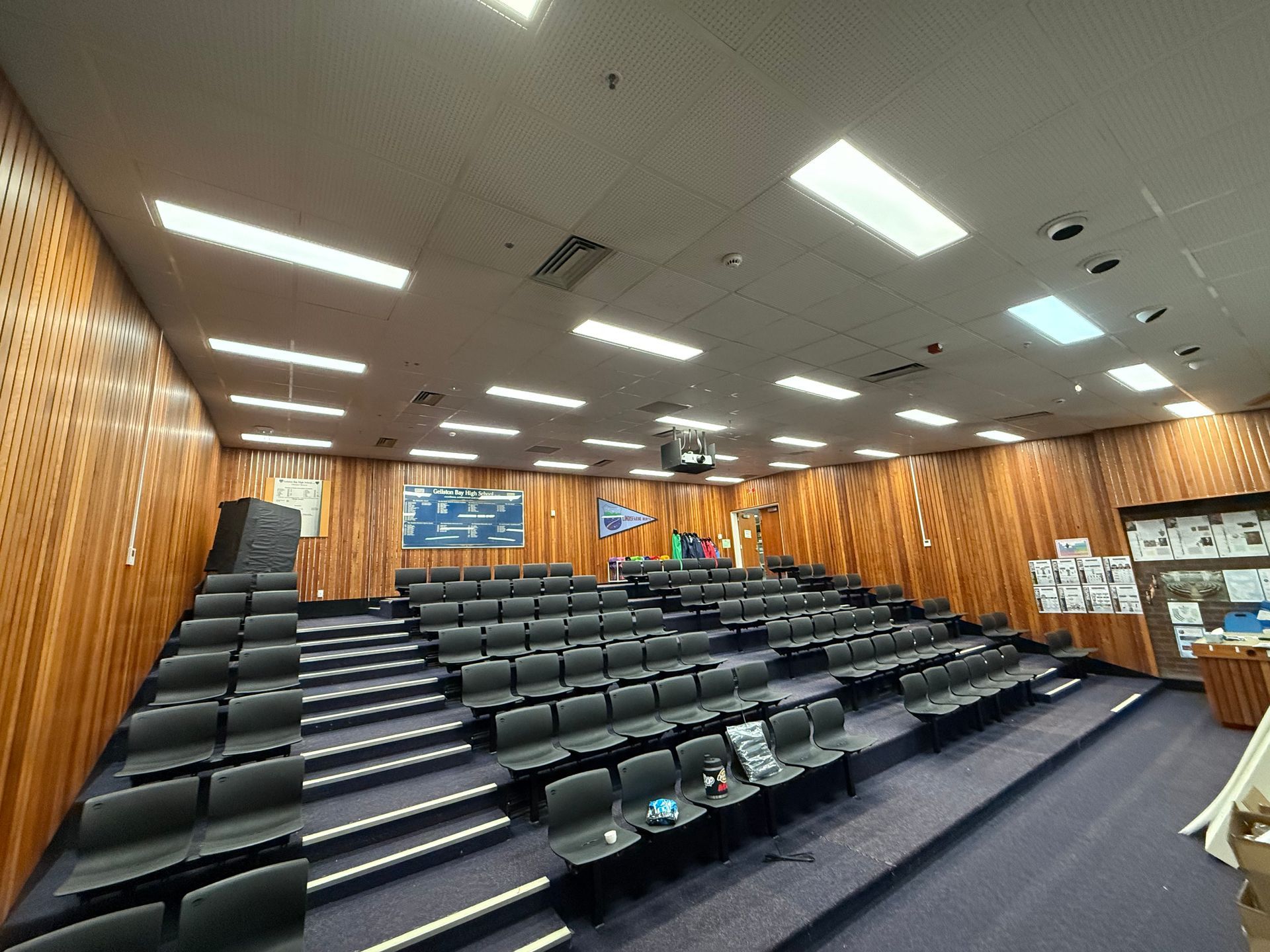 Wood-paneled lecture hall with tiered seating and bright ceiling lights; empty desks face the front stage.