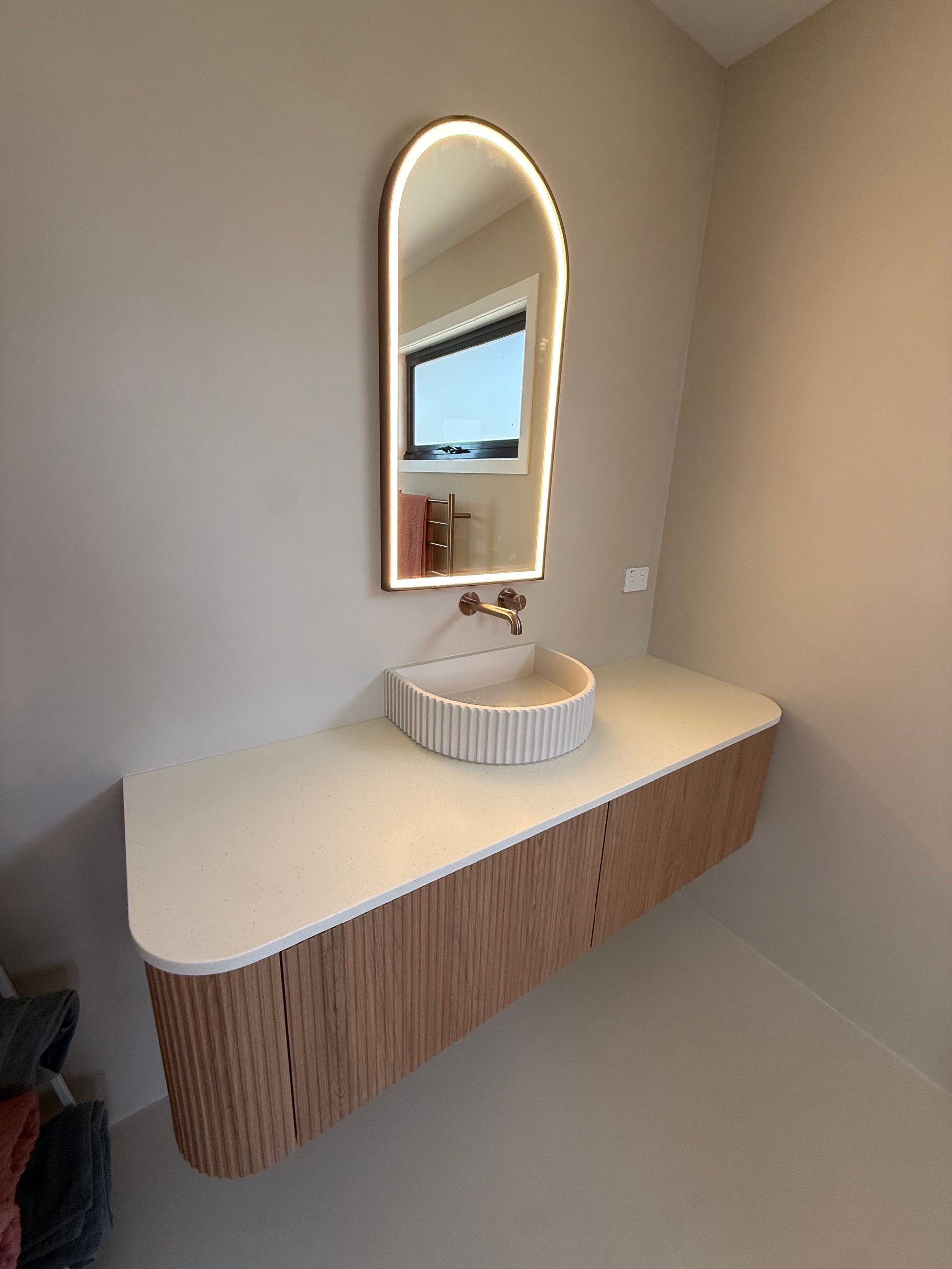 Modern bathroom vanity with cream countertop, striped wood base, round sink, and arched mirror.