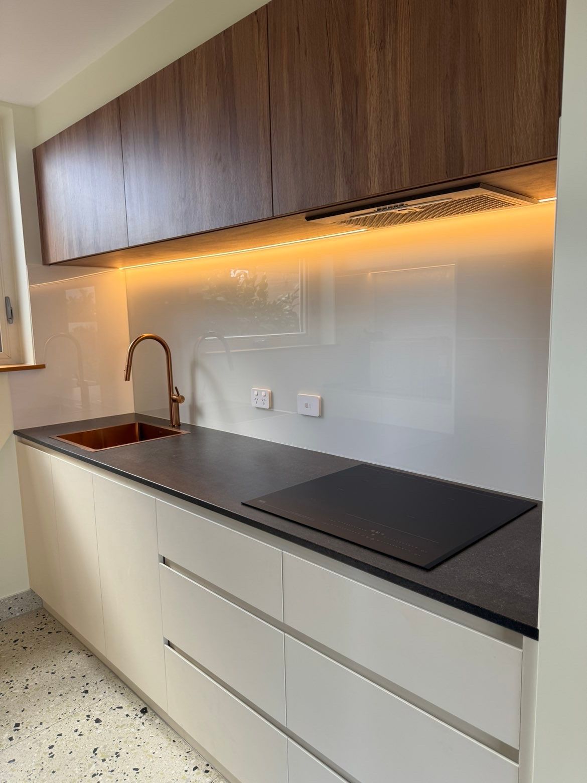 Modern kitchen counter with sink, black cooktop, white drawers, and wood cabinets with under-cabinet lighting