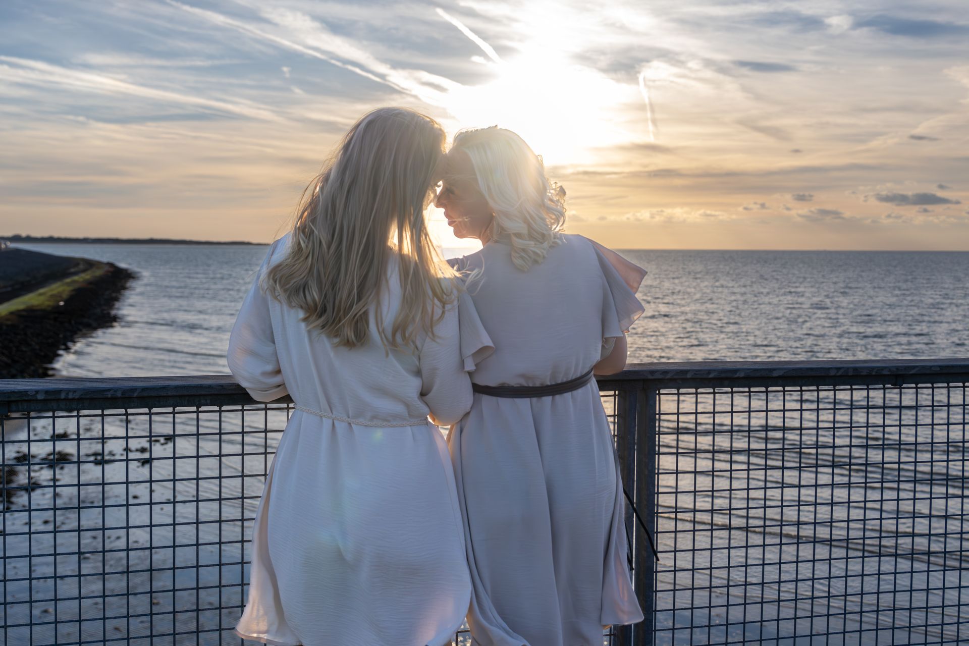Twee mensen in witte jurken staan bij zonsondergang aan zee en kijken over het water.
