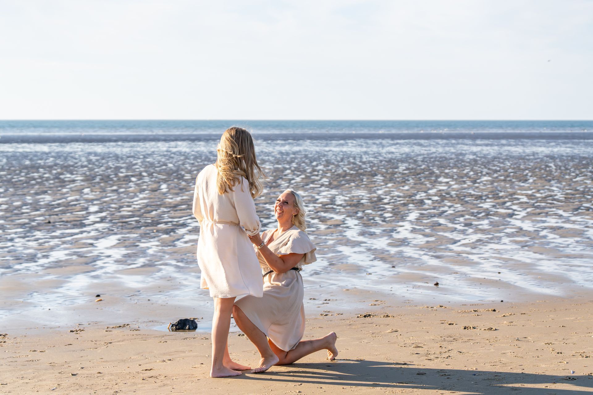 Twee mensen in witte kleding op een zandstrand aan het water, de een knielend en de hand van de ander vasthoudend.
