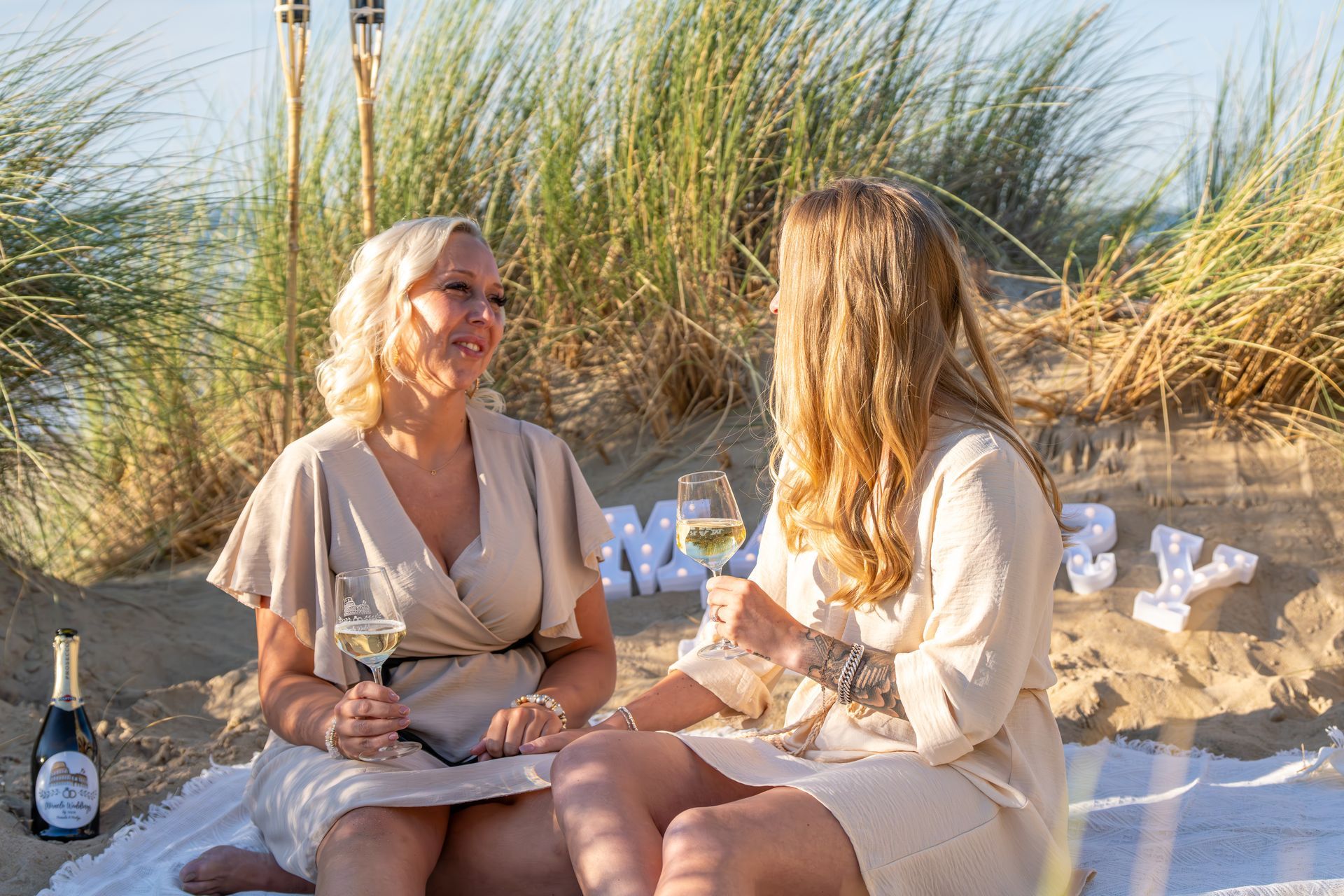 Twee vrouwen in lichte jurken zitten te kletsen op een zandstrand met een drankje en een fles wijn, met duinen op de achtergrond.