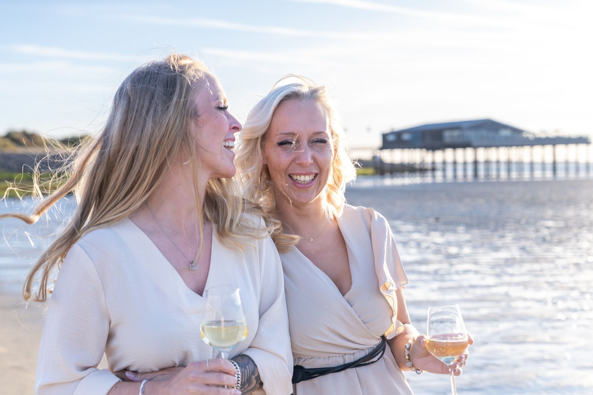 Twee vrouwen glimlachen en houden wijnglazen vast op een zonnig strand met een pier op de achtergrond.