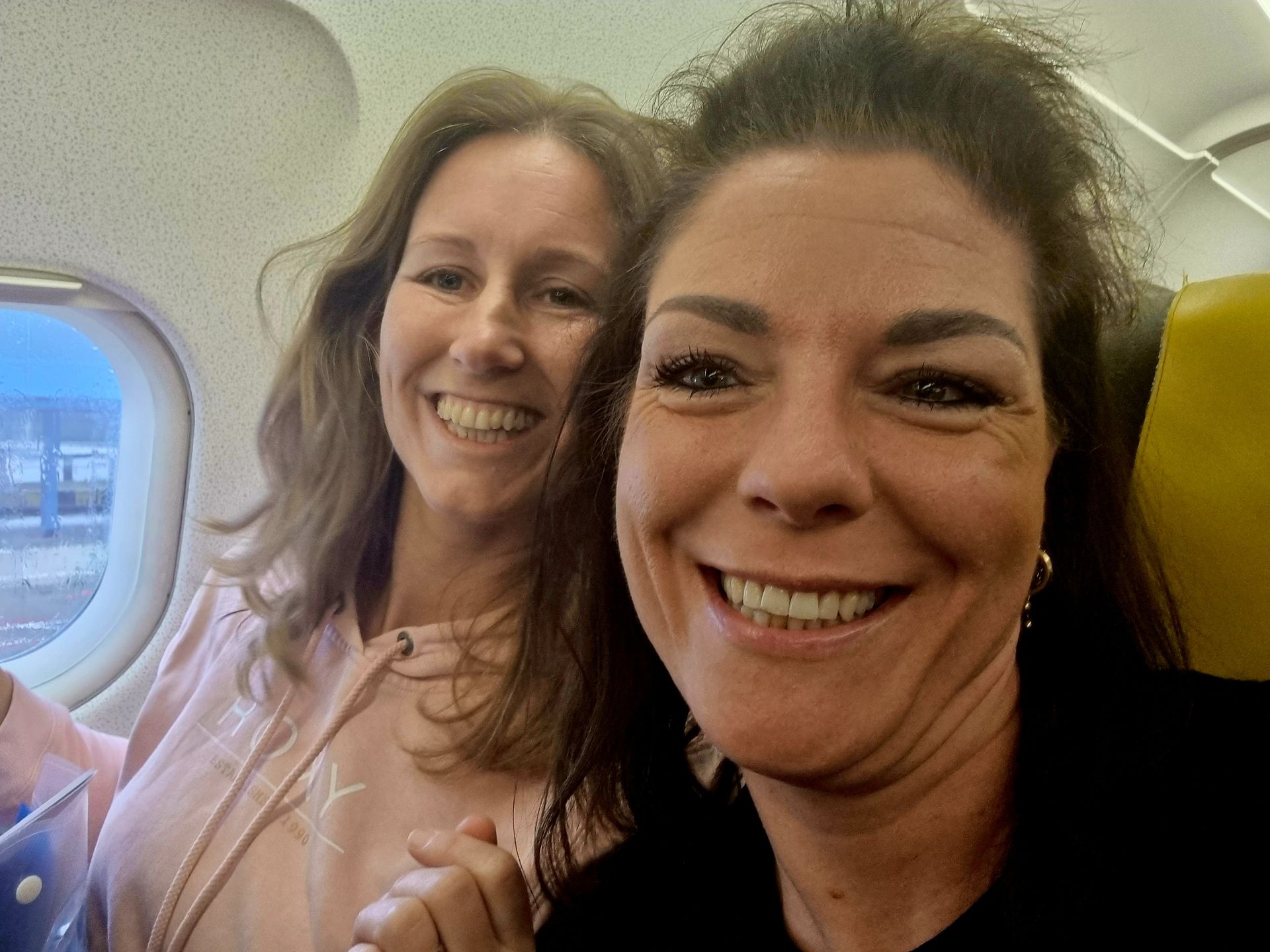 Twee lachende vrouwen maken een selfie in een vliegtuig, waarbij een raam en een gele hoofdsteun zichtbaar zijn.