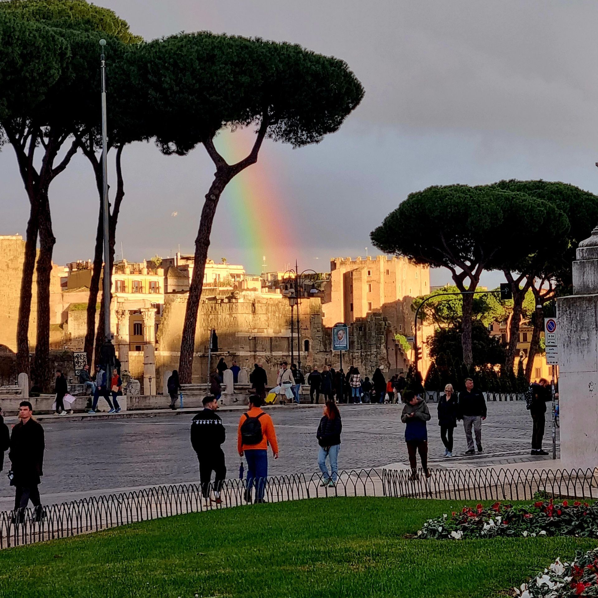 Een regenachtig plein met wandelende mensen, hoge parasoldennen en een regenboog boven historische gebouwen bij zonsondergang.