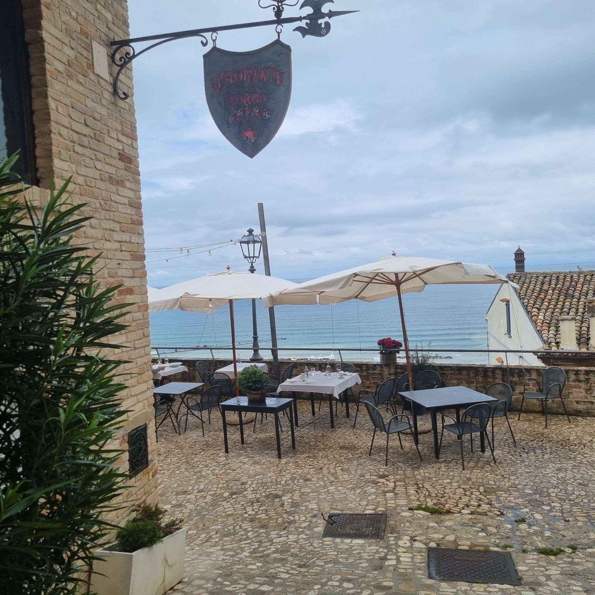 Terras van een café aan zee met witte parasols, tafels en stoelen op een geplaveid terras met uitzicht op de zee.