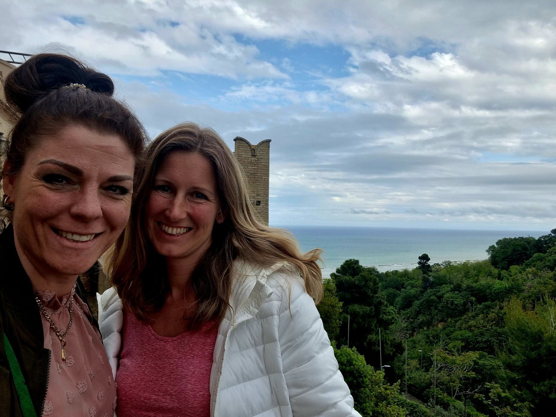 Twee lachende vrouwen maken een selfie in de buitenlucht met de oceaan en een bewolkte hemel op de achtergrond.