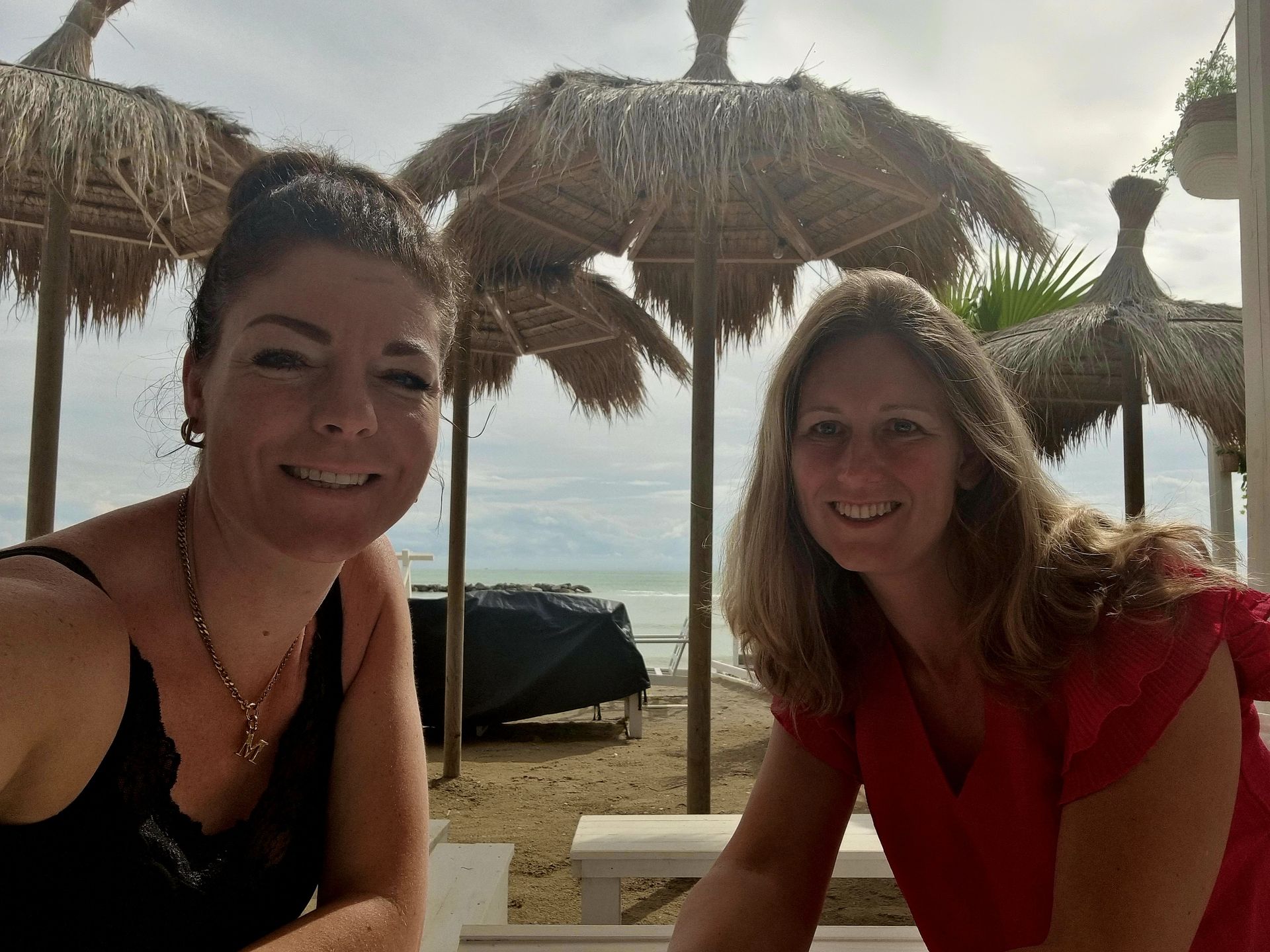 Twee vrouwen zitten onder rieten parasols op een zandstrand en lachen naar de camera.