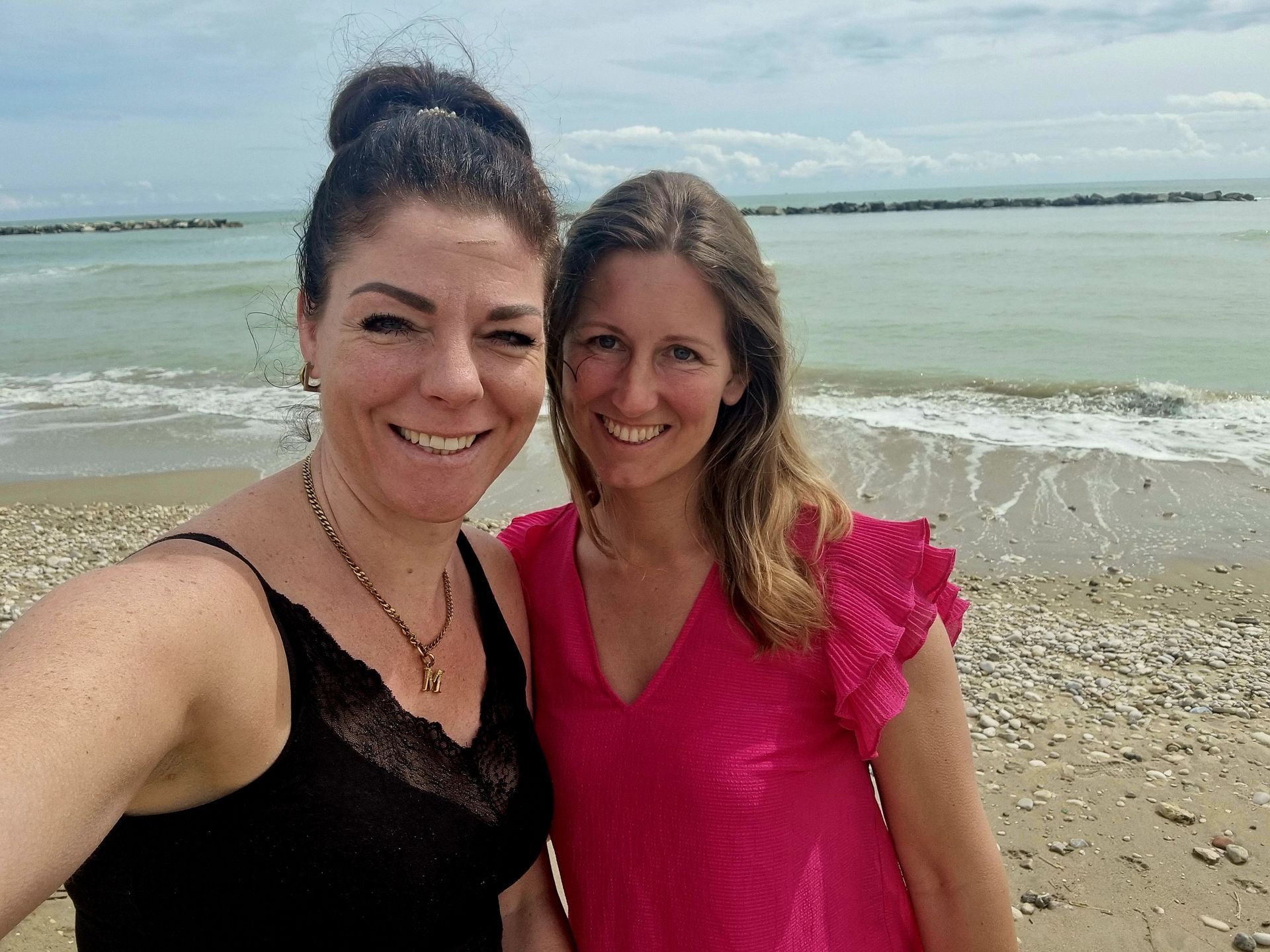 Twee vrouwen poseren lachend voor een selfie op het strand, met de golven en de kustlijn op de achtergrond.