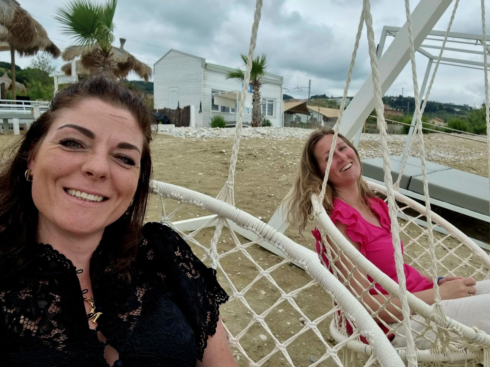 Twee vrouwen liggen ontspannen in een witte hangmat op het strand, glimlachend met palmbomen en zand op de achtergrond.