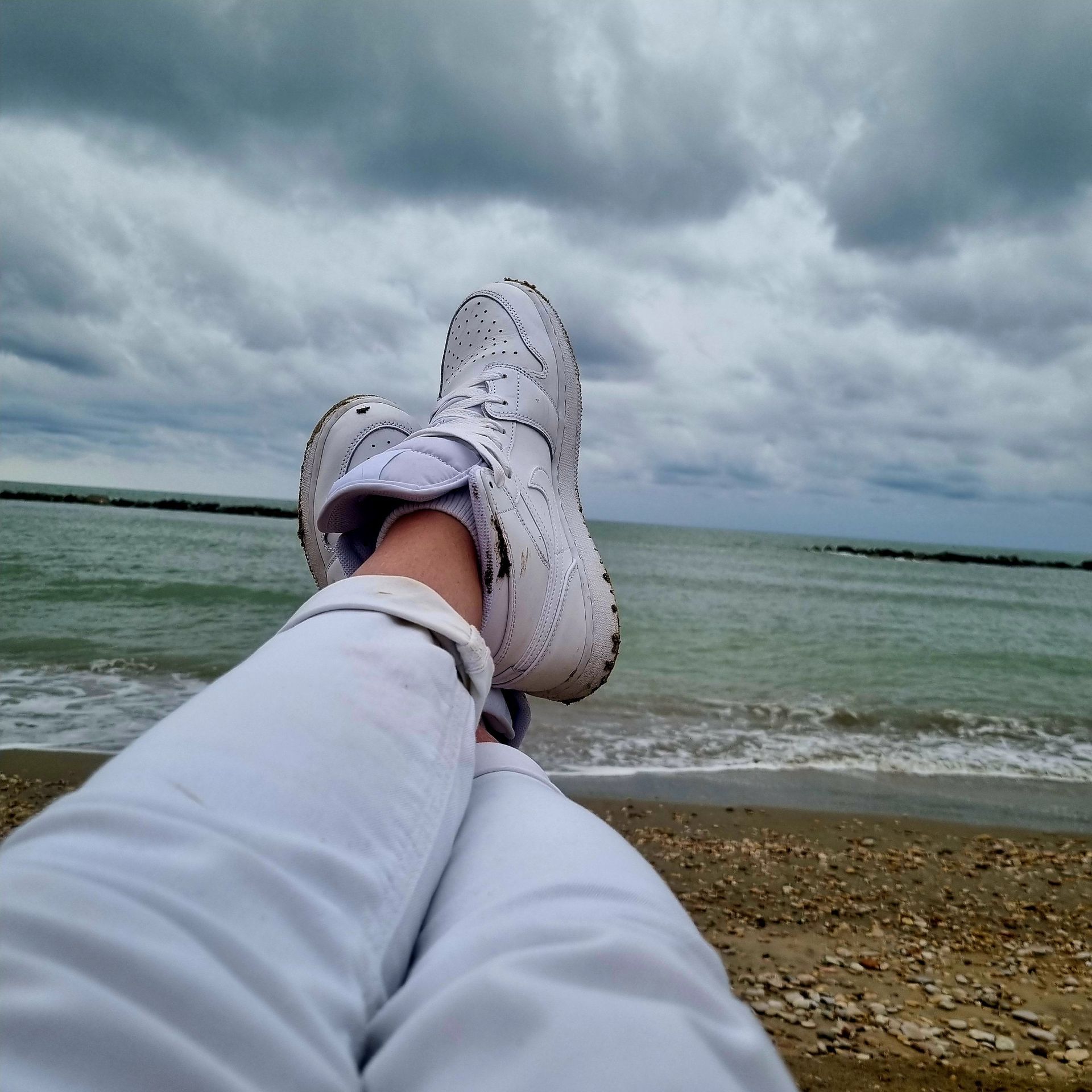 Voeten in witte sneakers uitgestrekt op een strand onder een bewolkte hemel, met uitzicht op het water.