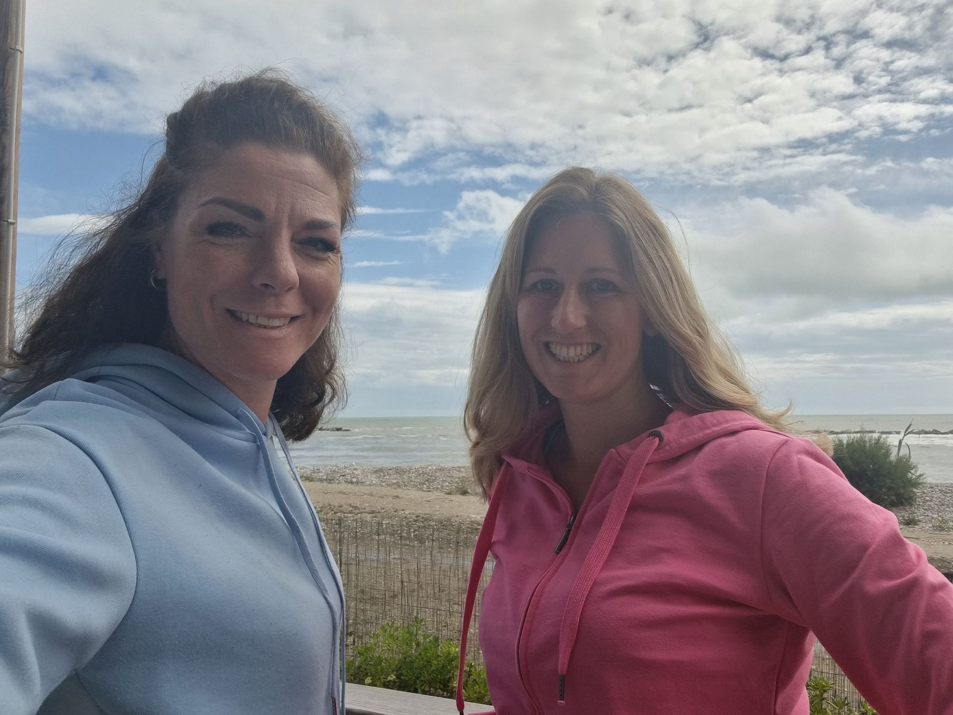 Twee vrouwen glimlachen buiten op het strand, de ene in een blauwe hoodie en de andere in een roze hoodie.