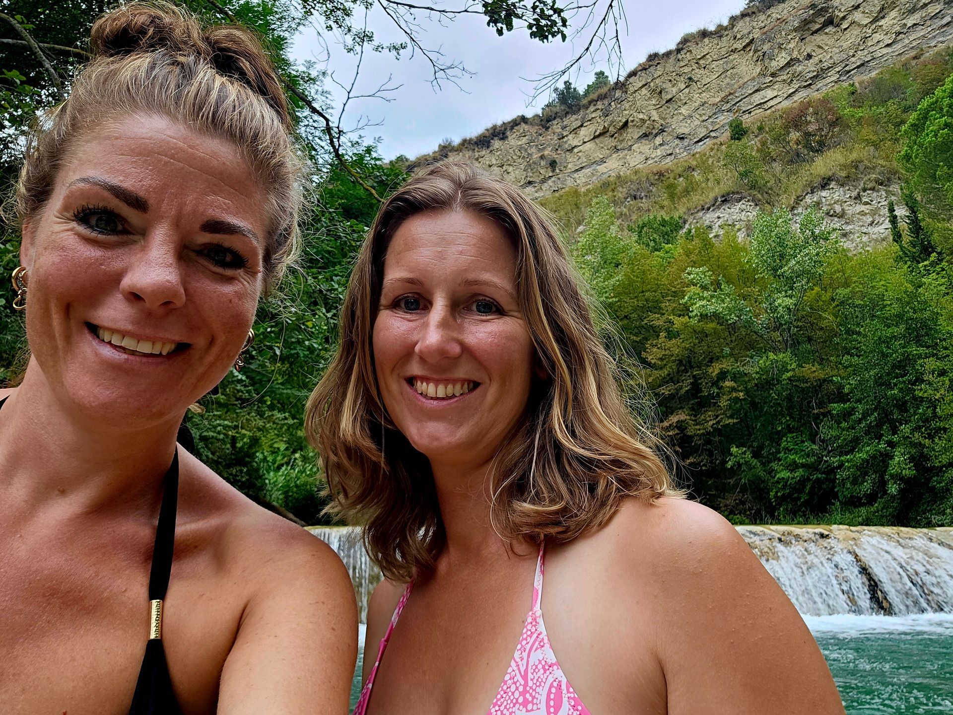 Twee lachende vrouwen poseren buiten met bergen en groen op de achtergrond.