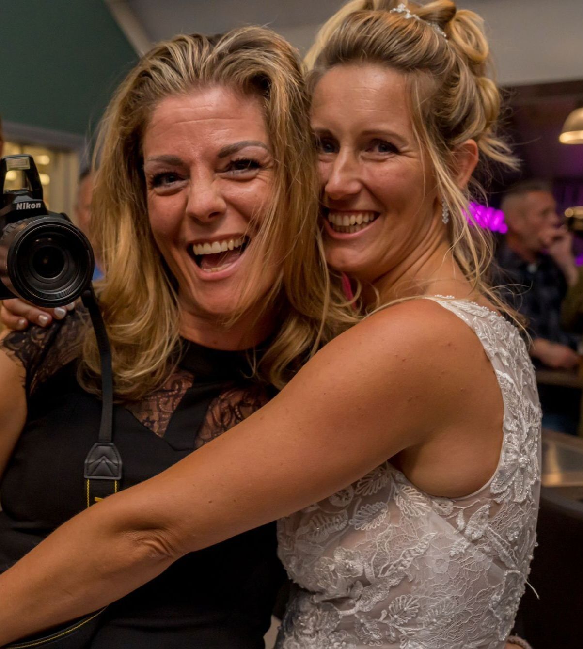 Twee lachende mensen omhelzen elkaar tijdens een evenement binnenshuis, een van hen houdt een camera vast, beiden in elegante jurken.