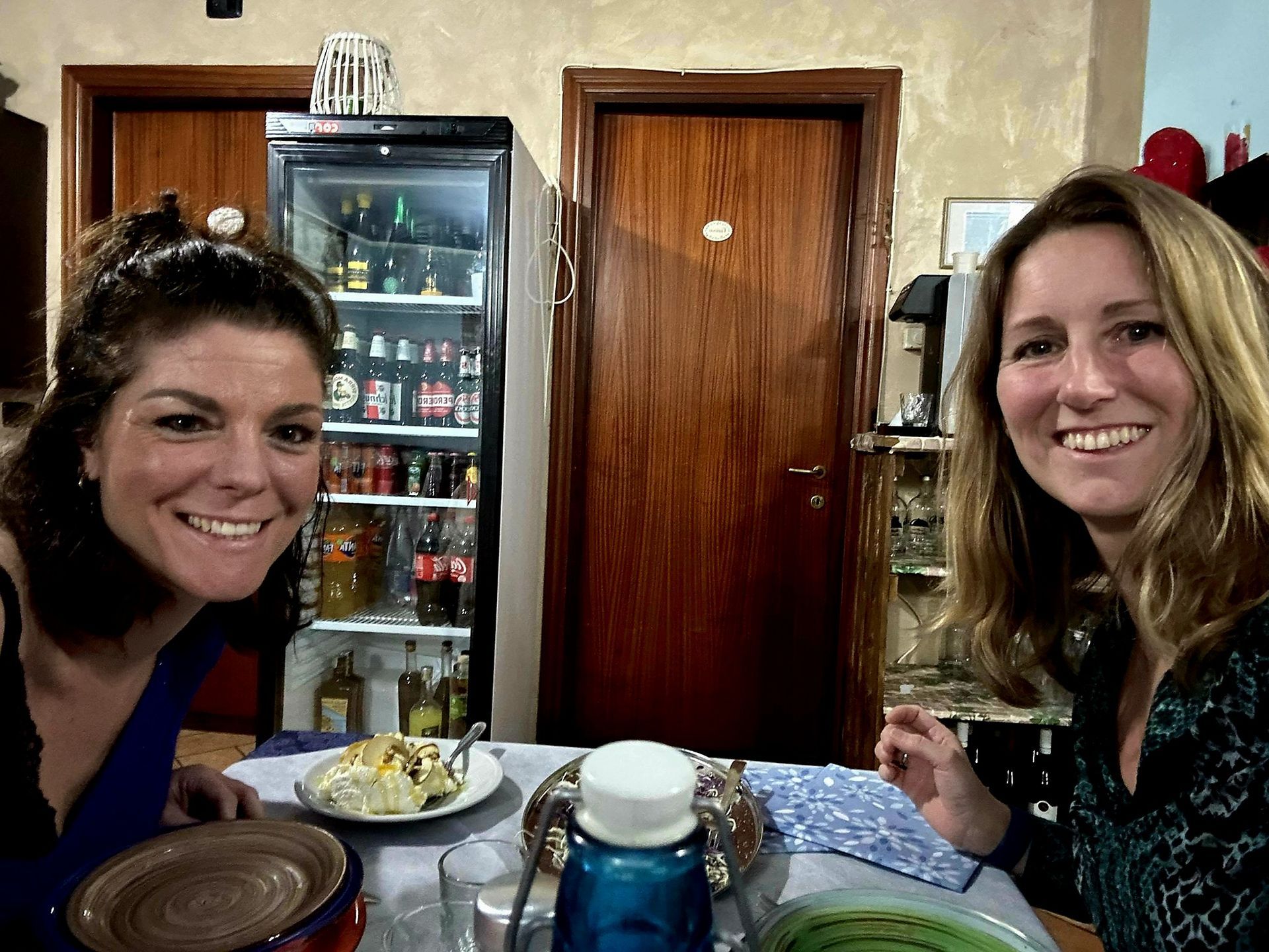 Twee vrouwen glimlachen aan een restauranttafel met drankjes en dessert voor zich.
