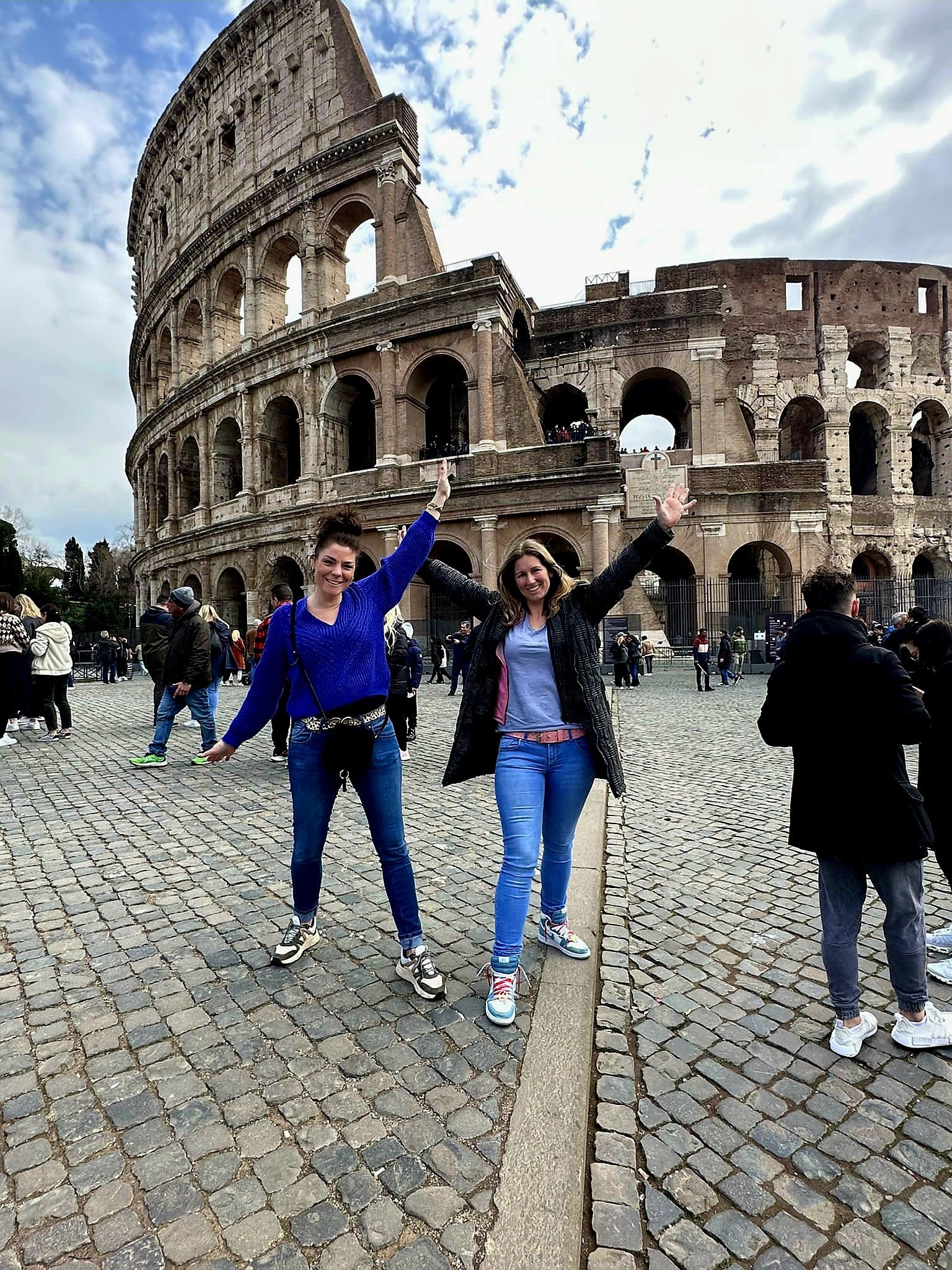 Twee mensen poseren voor het Colosseum in Rome, één van hen met een arm omhoog.
