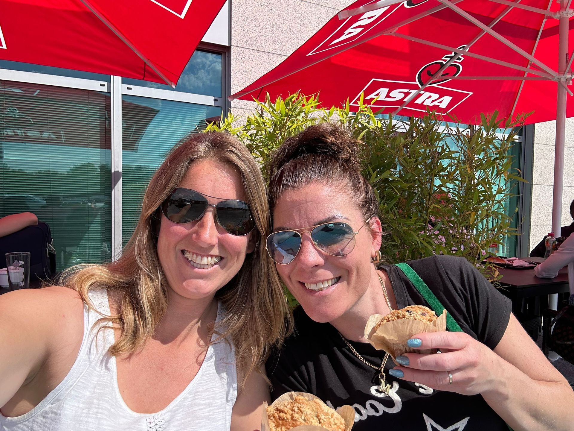 Twee lachende mensen onder rode parasols bij een terrasje, waarvan één een gebakje en een drankje vasthoudt.