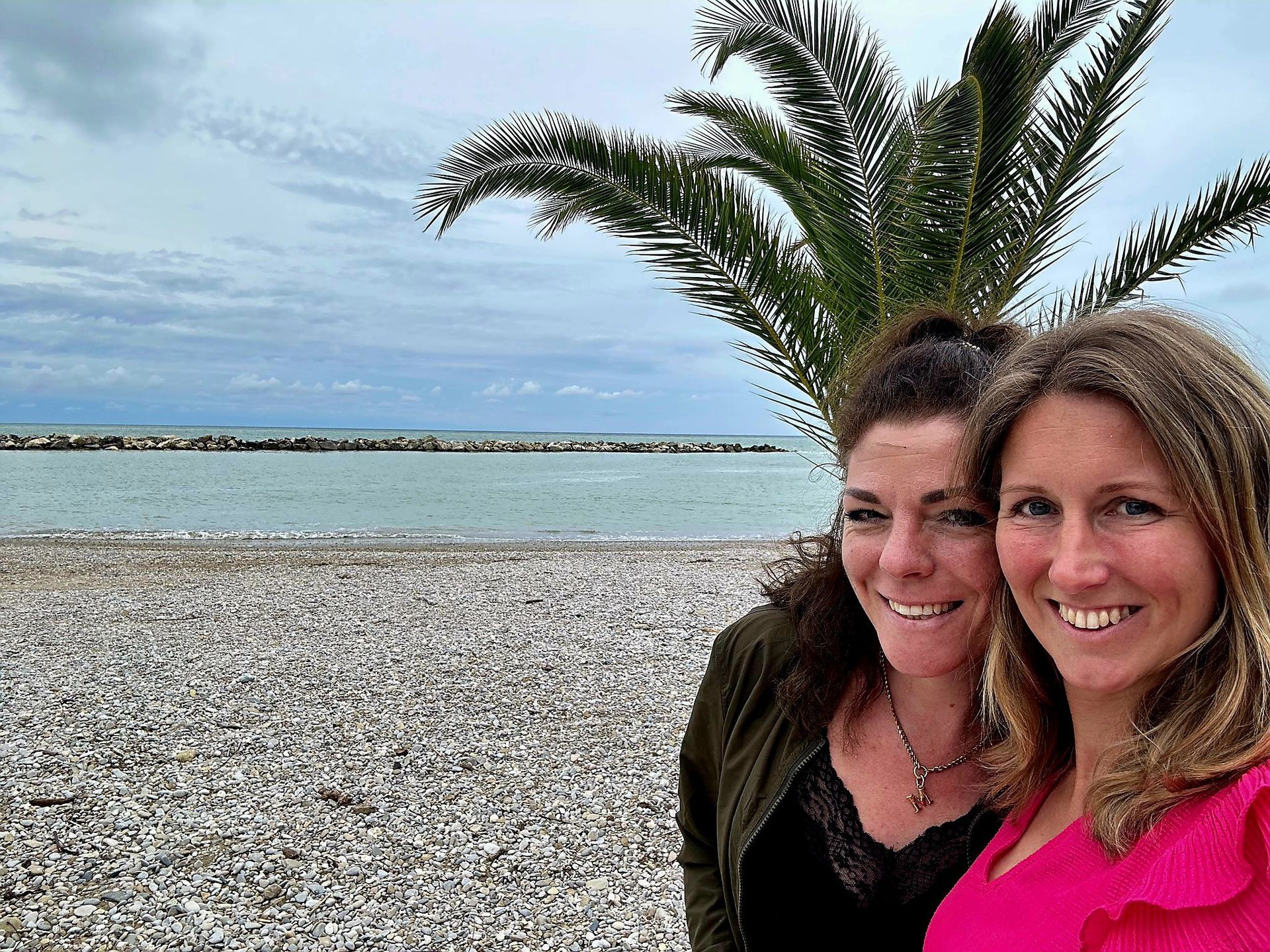 Twee mensen glimlachen op een kiezelstrand met een palmboom en een kalme blauwe zee op de achtergrond.