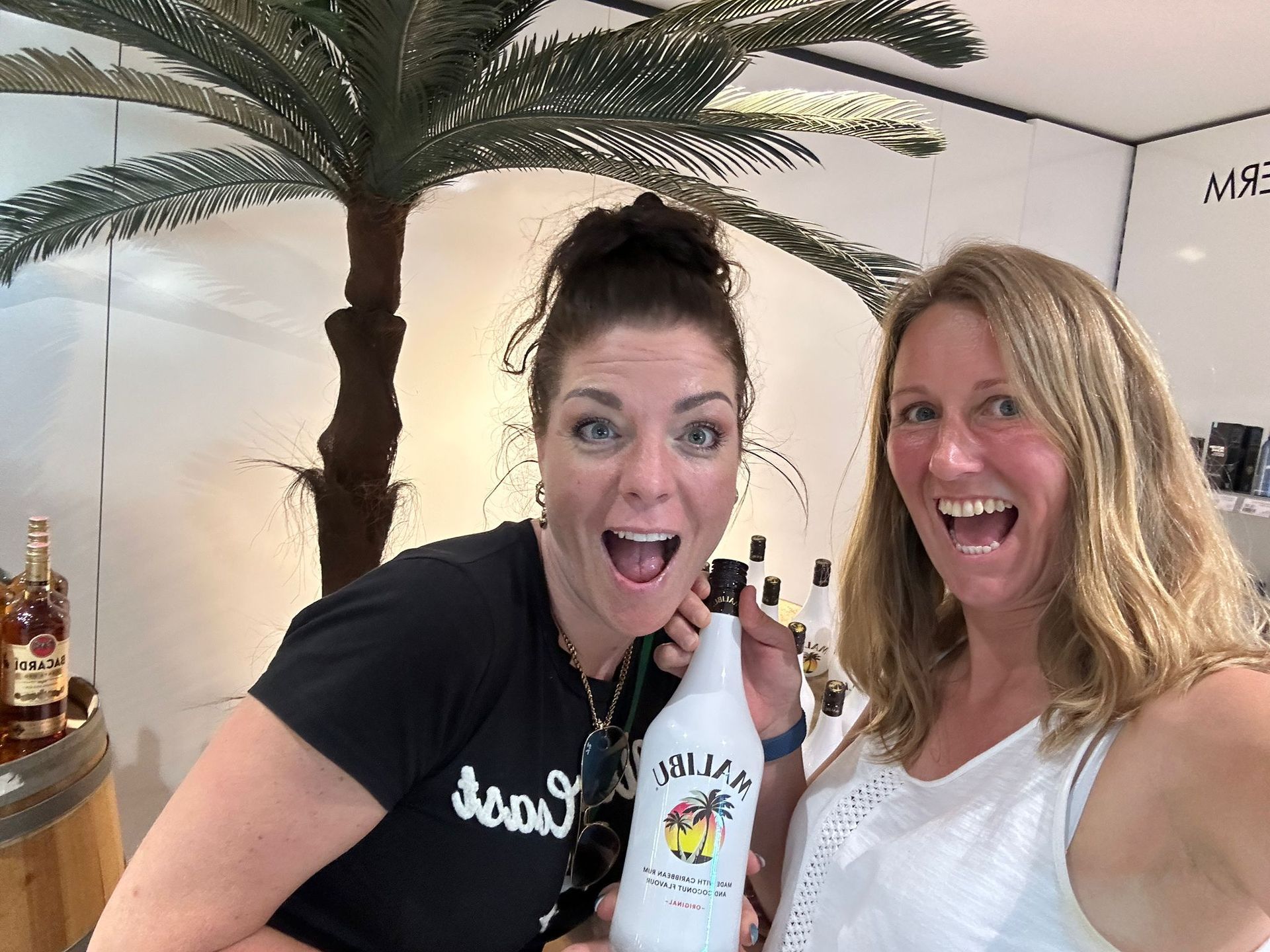 Twee lachende vrouwen poseren binnen met een witte fles en palmboomdecoratie op de achtergrond.
