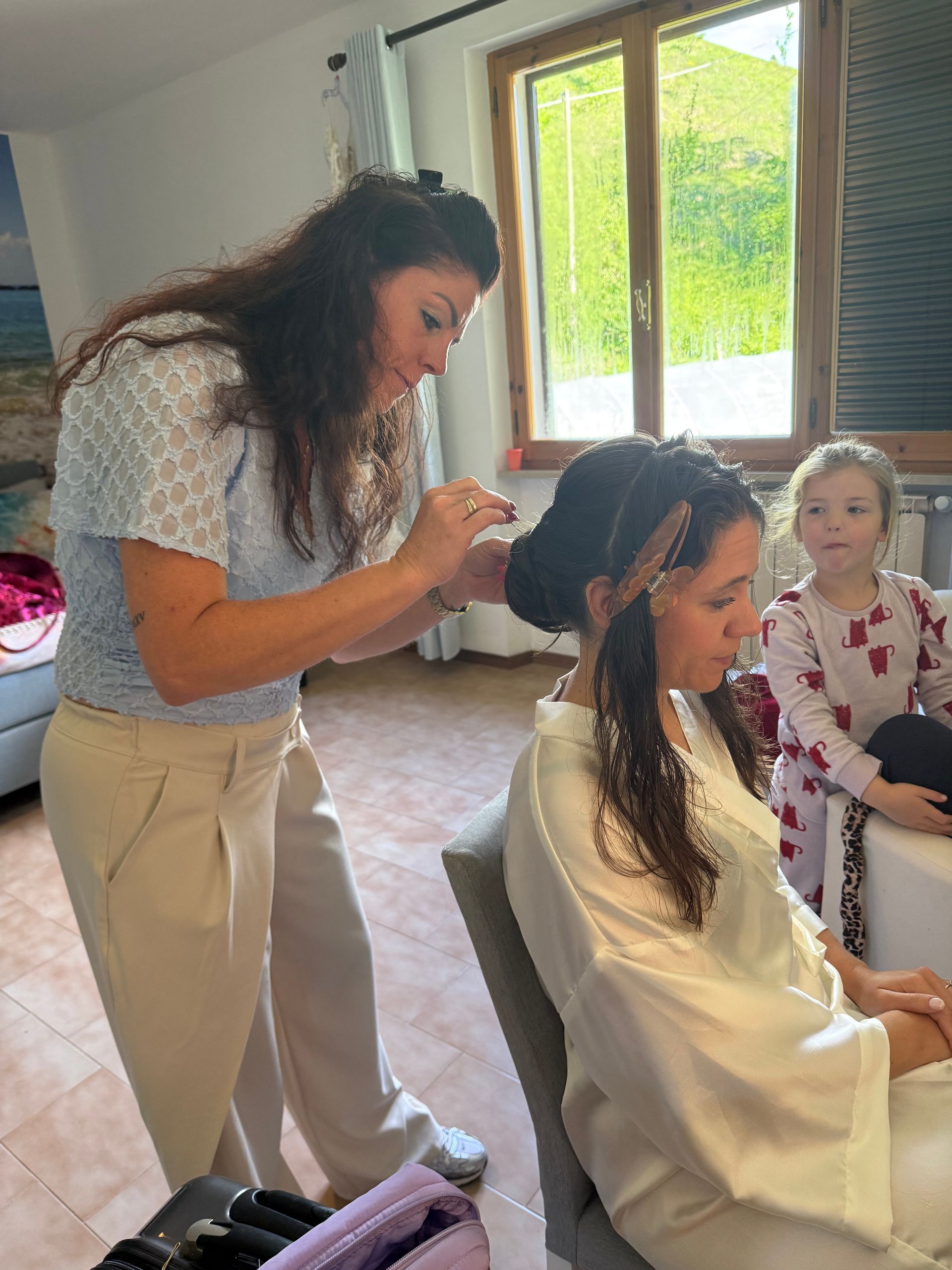 Een haarstyliste vlecht het haar van een zittende klant in een lichte salon, terwijl een andere klant in de buurt wacht.