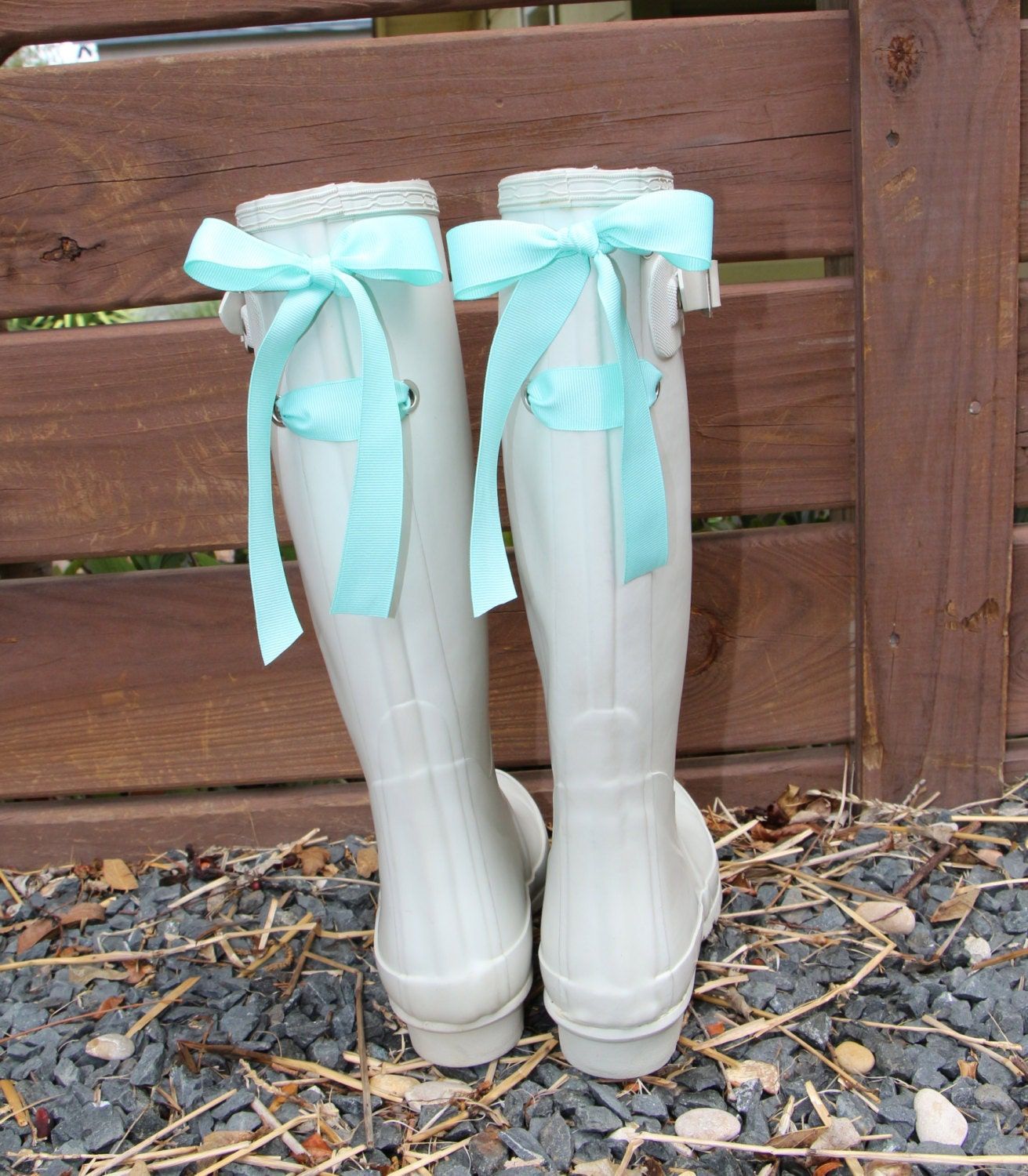 Witte regenlaarzen met aquakleurige strikken staan op grind voor een houten hek.
