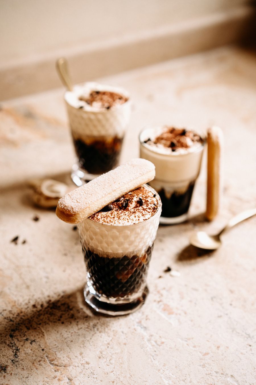 Drie gelaagde koffiedranken, afgetopt met slagroom en cacao, op een beige aanrecht.