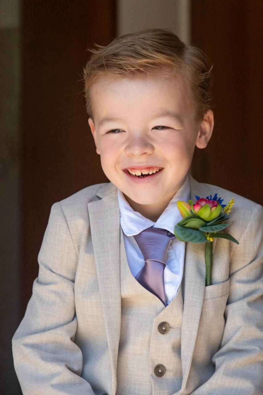 Een lachend kind in een lichtgrijs pak met een paarse stropdas en boutonnière, staand tegen een donkere achtergrond.