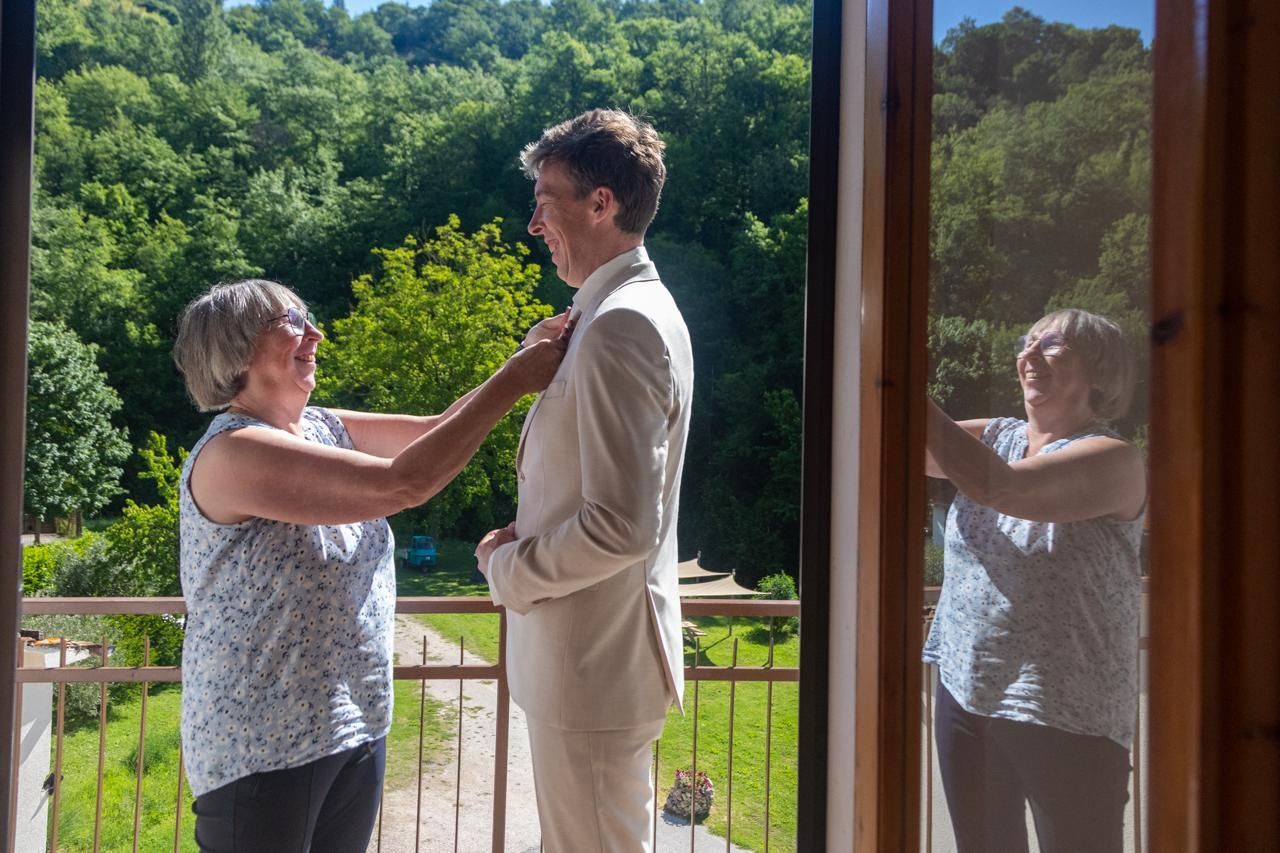 Een vrouw maakt de kraag van een man recht op een zonnig balkon, met groene bomen op de achtergrond.