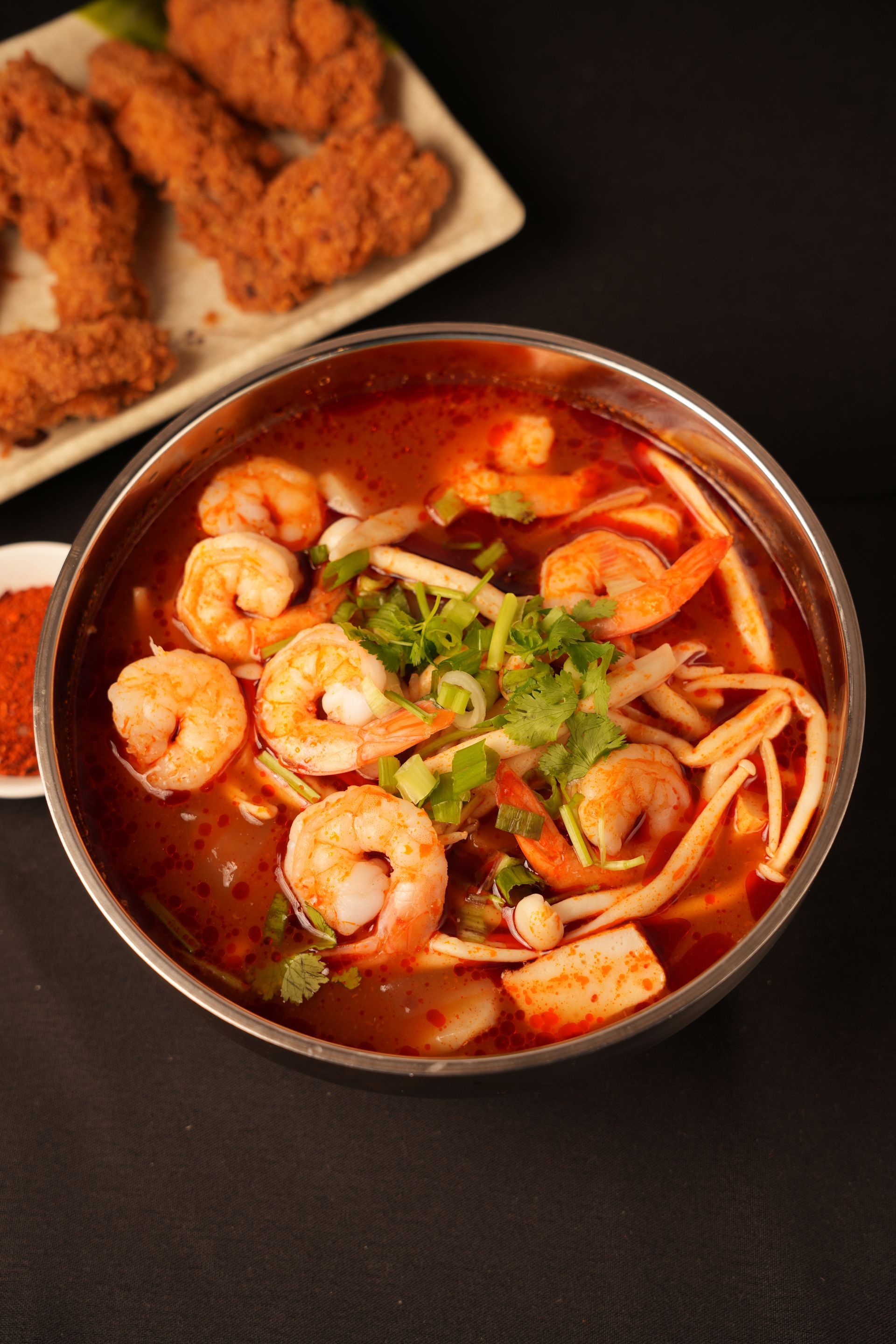 Bowl of red shrimp noodle soup with herbs, beside fried chicken pieces on a dark table