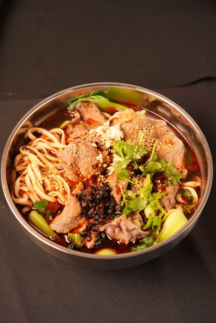 Bowl of noodles topped with sliced meat and greens on a dark table