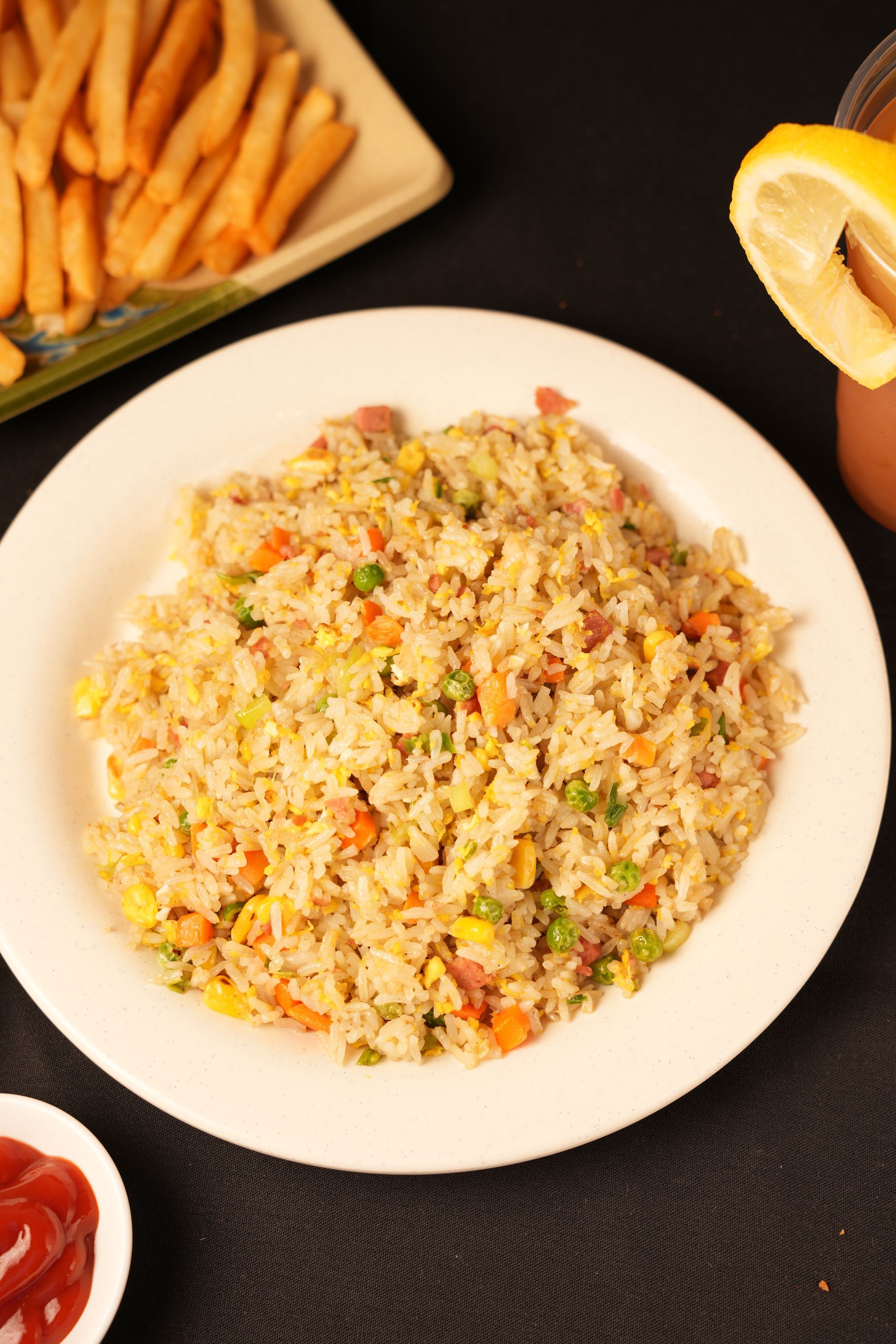 Plated fried rice with vegetables, served with fries, ketchup, and lemon on a black table.