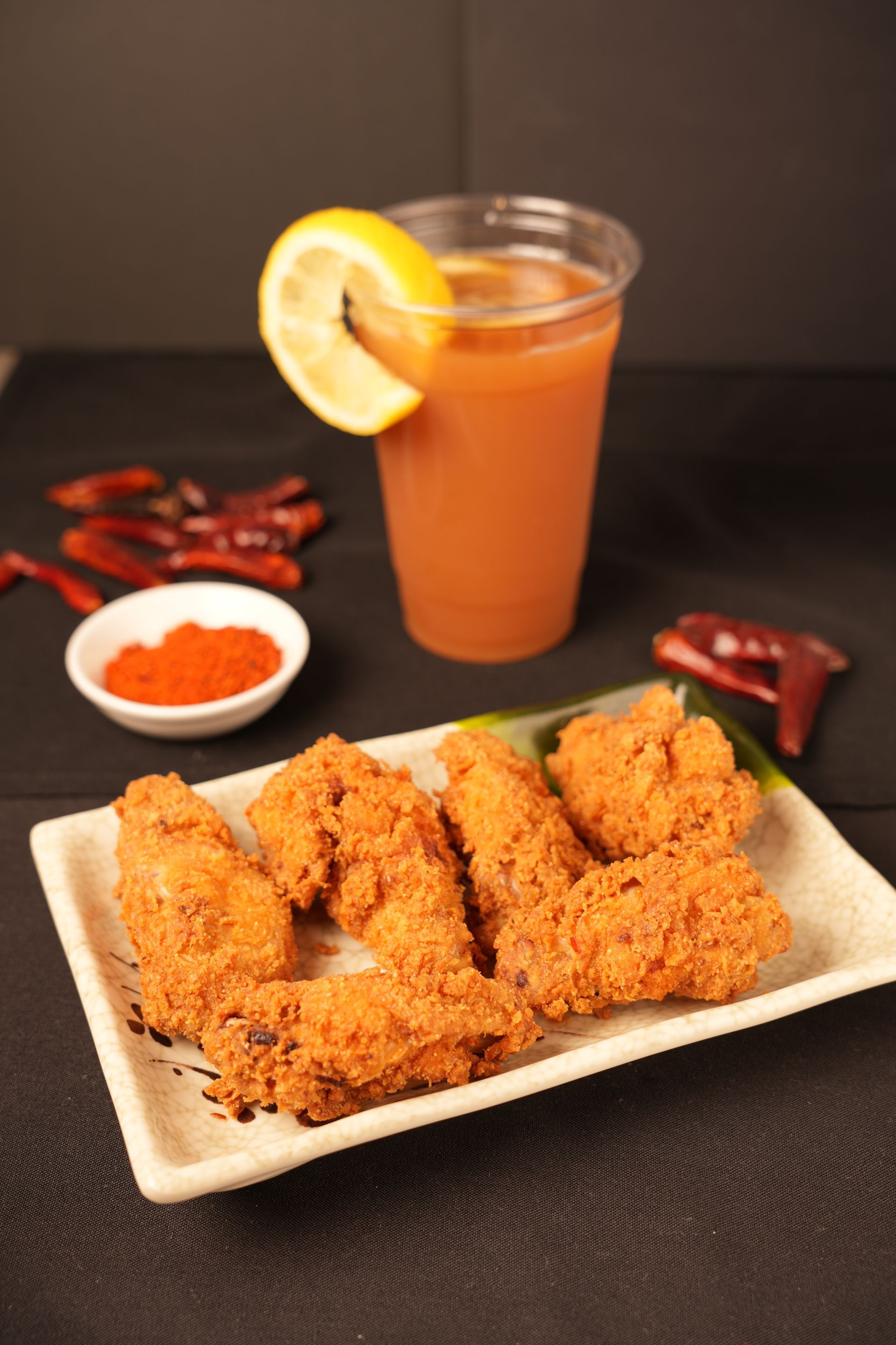 Fried chicken strips on a plate with orange dipping sauce, a lemon-garnished drink, and chili peppers