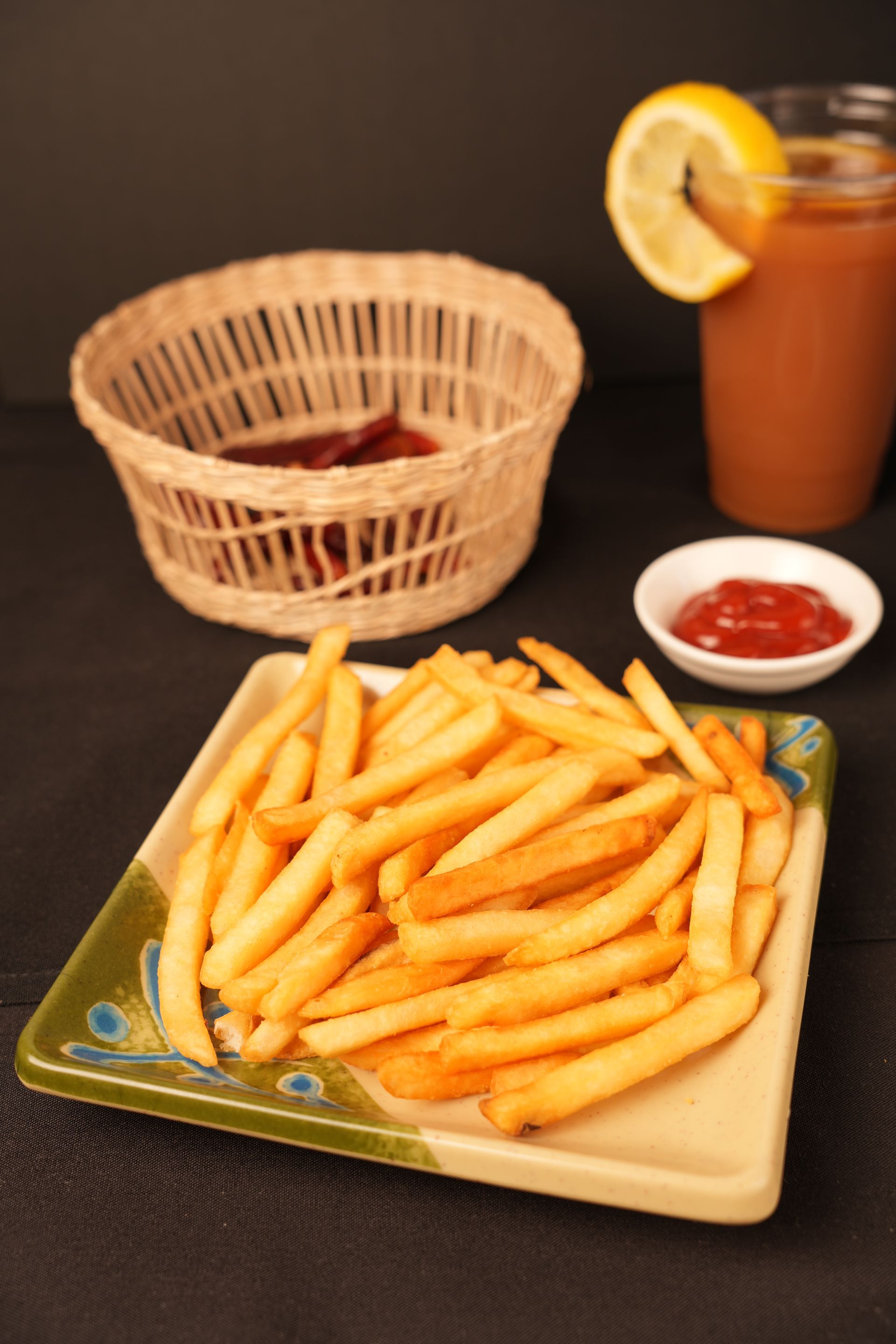 French fries on a plate with ketchup, a basket, and a drink with a lemon slice