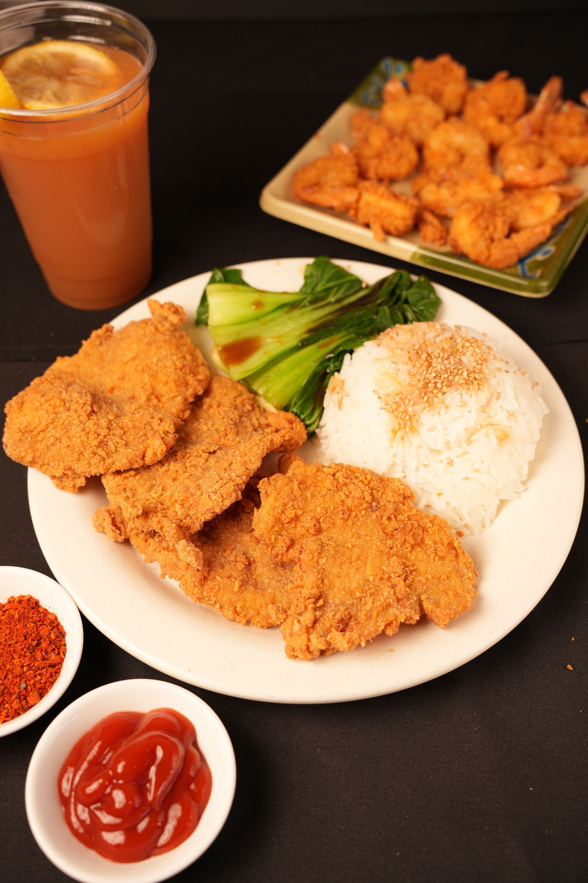 Fried chicken cutlets with rice, sauces, and iced tea on a black table, plus a side of chicken bites